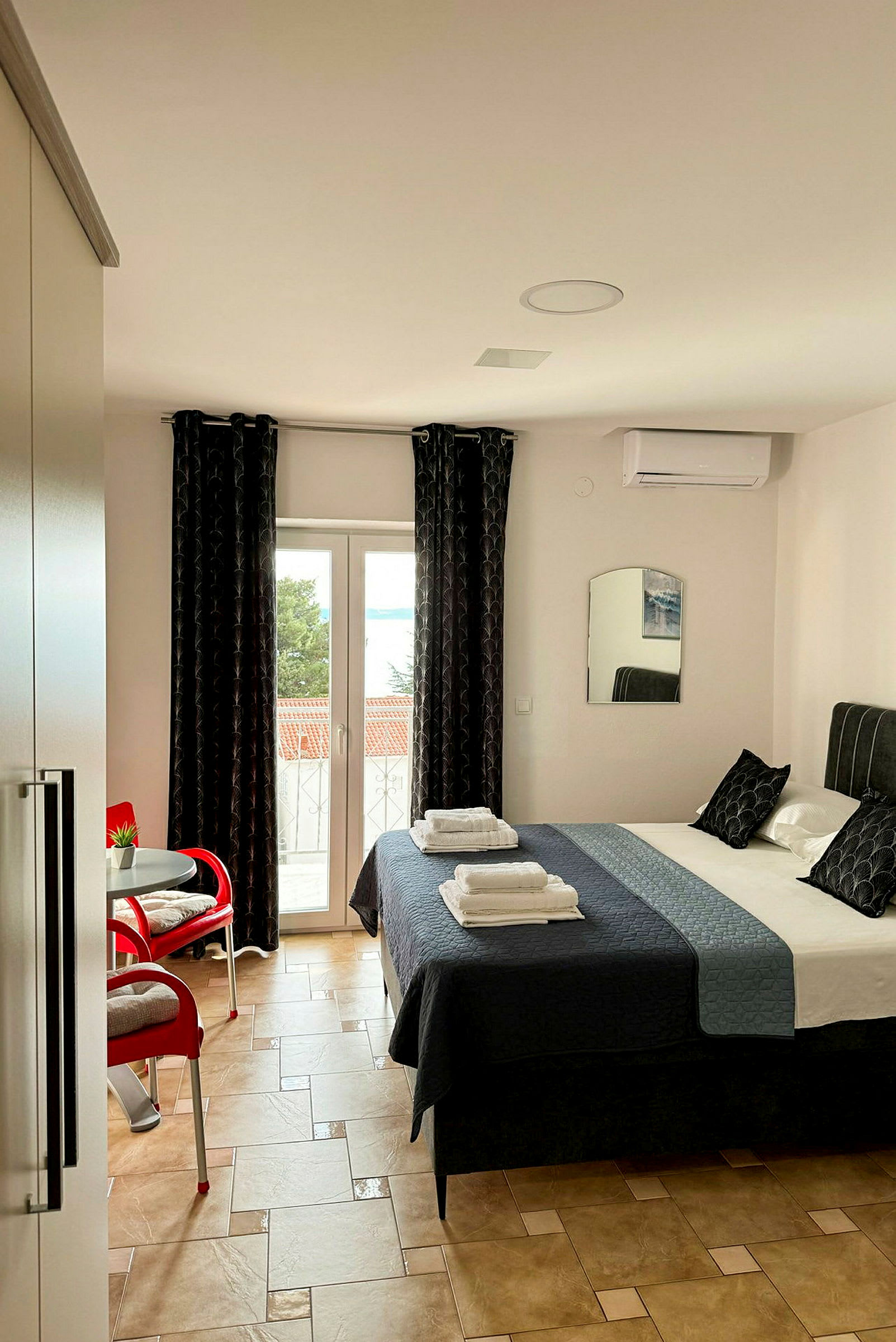Ett hotellrum med en dubbelsäng, två uppsättningar handdukar på sängen, ett litet bord med en blomma och två röda stolar samt glasdörrar som leder ut till en balkong. (AI-genererad text)