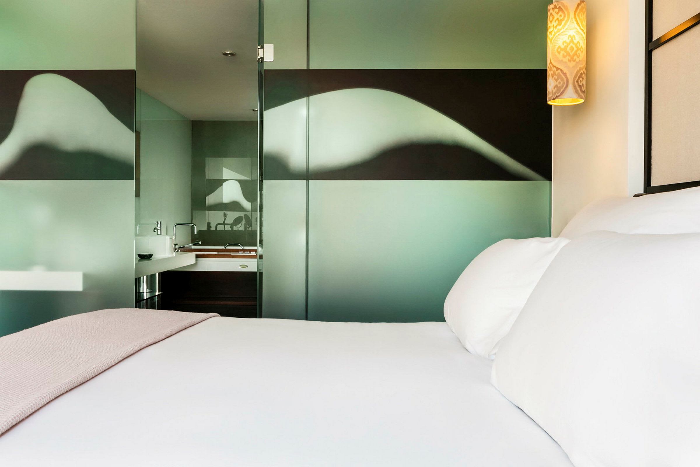 Ett modernt hotellrum visar en säng med vita lakan och en rosa filt, samt ett glasparti med mönster som avskiljer badrummet. (AI-genererad text)