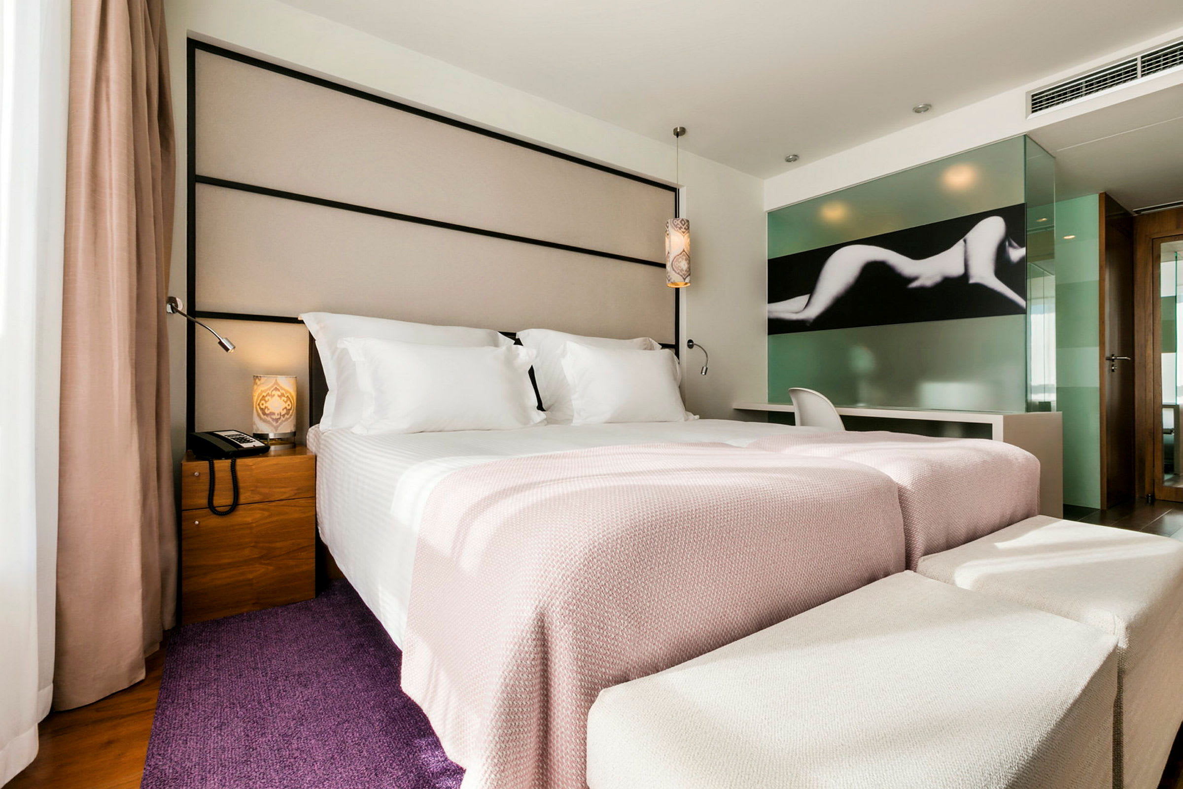 Ett modernt hotellrum med en bred säng bäddad med vita och ljusrosa sängkläder. Det finns en stol och ett skrivbord vid en glasvägg med konstmotiv. (AI-genererad text)