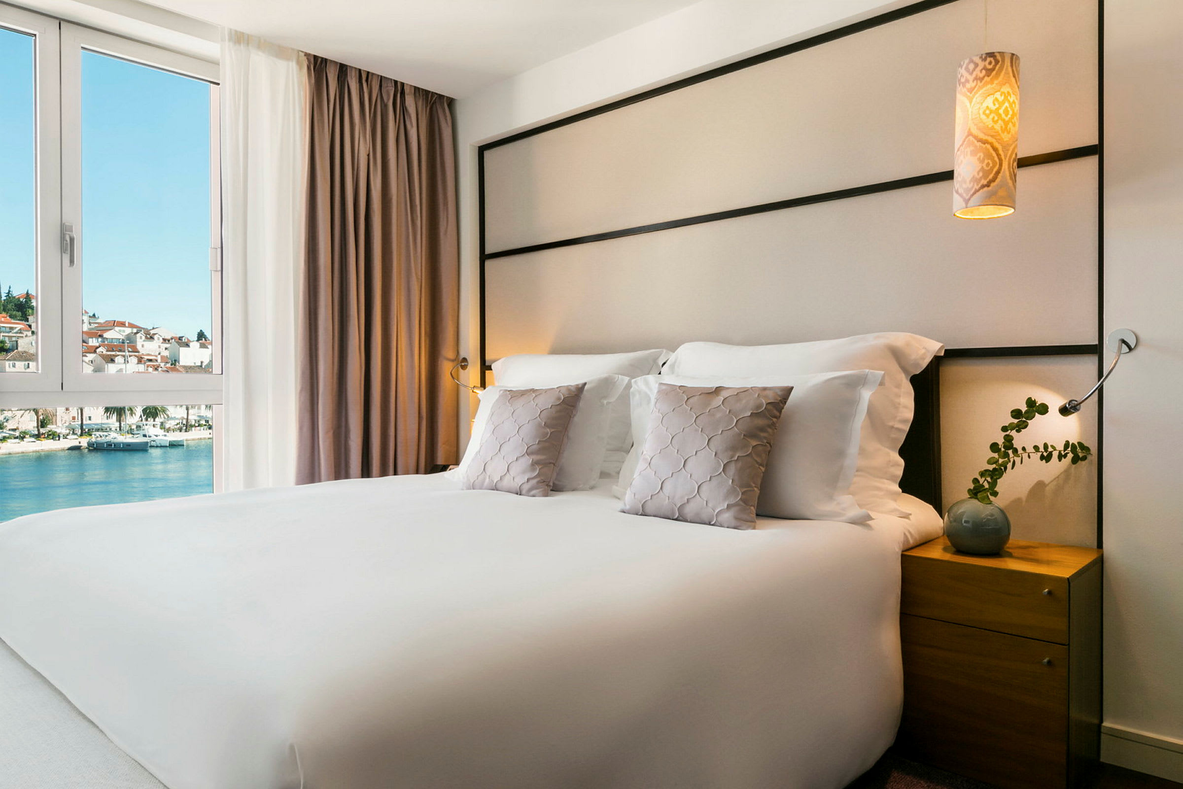 Hotellrum med en dubbelsäng, prydnadskuddar och ett nattduksbord. Utsikt över vatten och byggnader genom fönstret. (AI-genererad text)