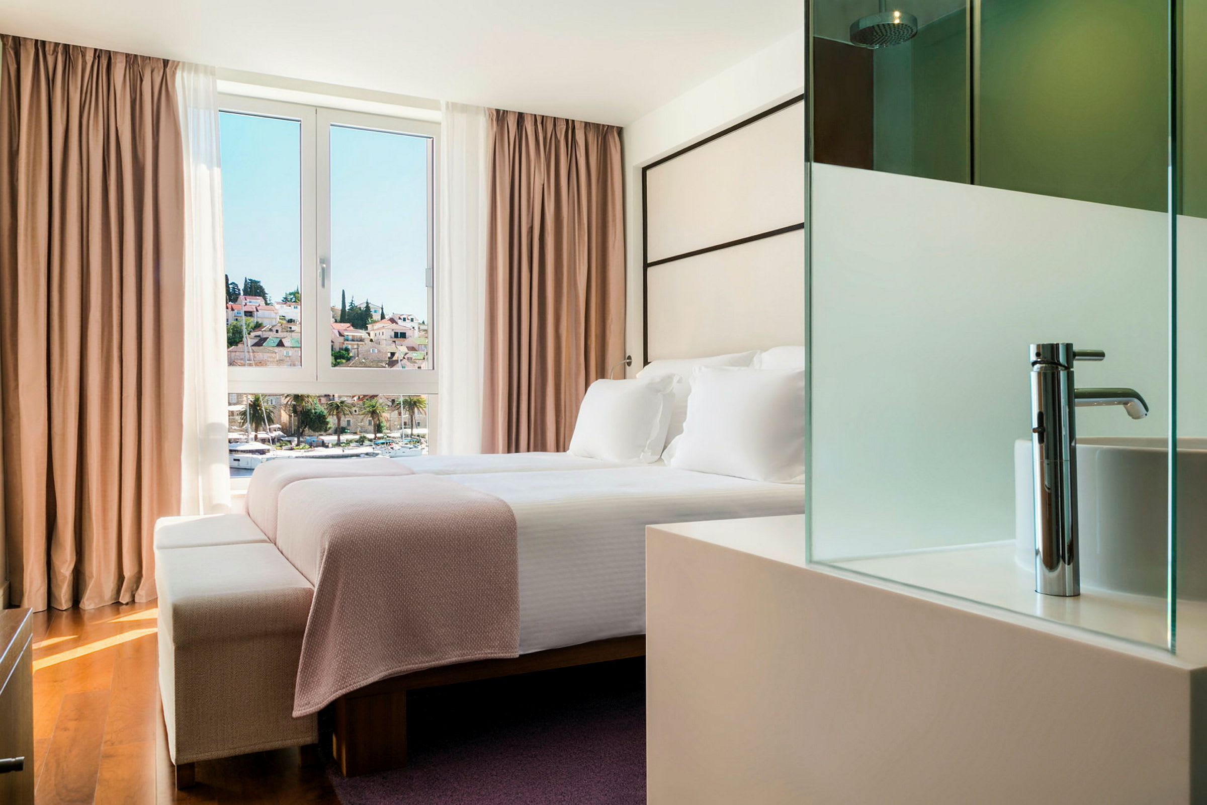Ett hotellrum med en dubbelsäng placerad framför ett stort fönster med utsikt över en stad. Sängen är bäddad med vita lakan och en rosa pläd. (AI-genererad text)