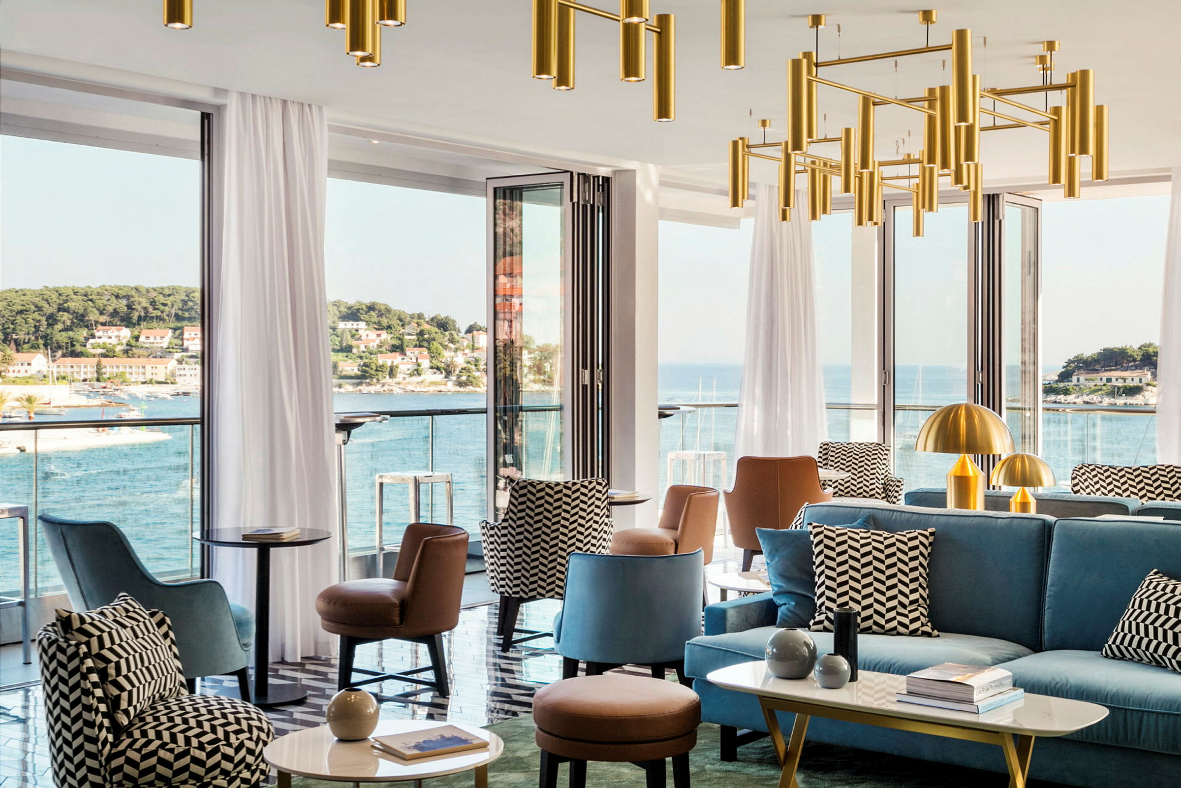 En modern lounge med sittmöbler och dekorativa ljuskronor, omgiven av stora fönster som erbjuder utsikt över en kuststad och havet. (AI-genererad text)