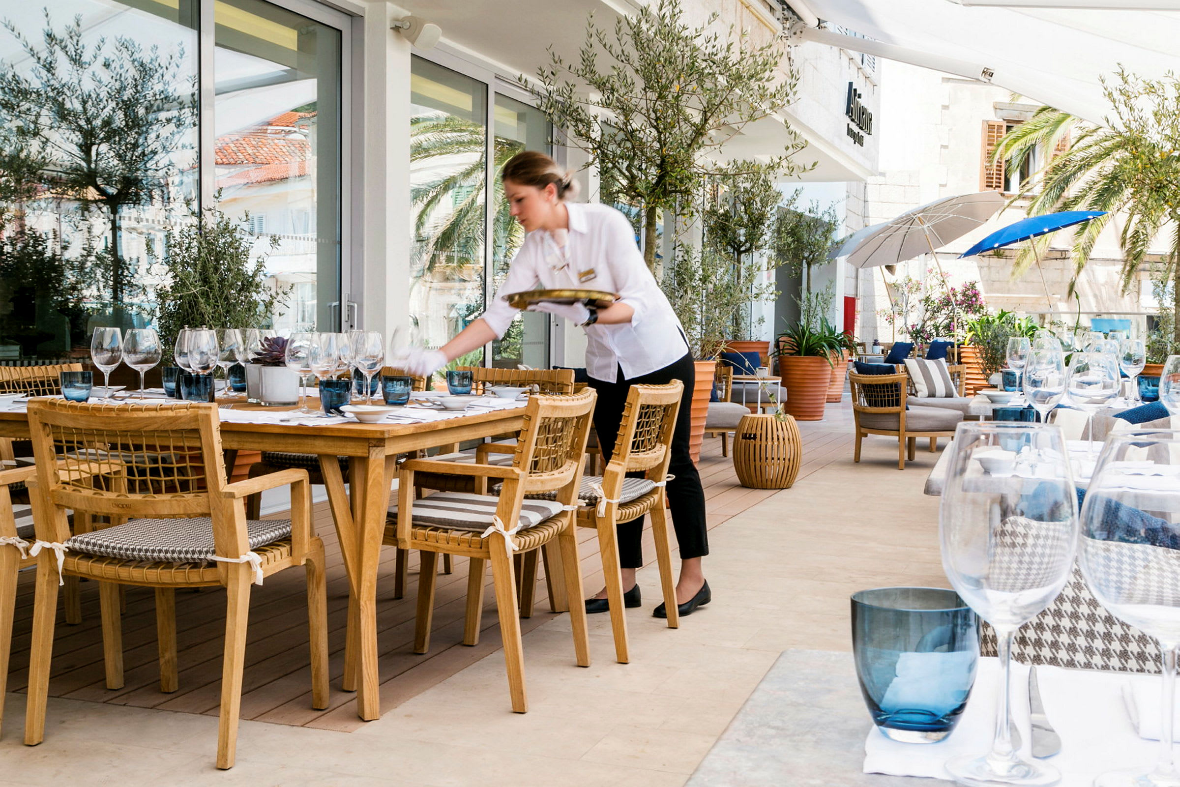 En utomhusrestaurang med träbord och stolar samt dukningar innehållandes glas och porcelän. Fina omgivningar med växter och parasoller. (AI-genererad text)
