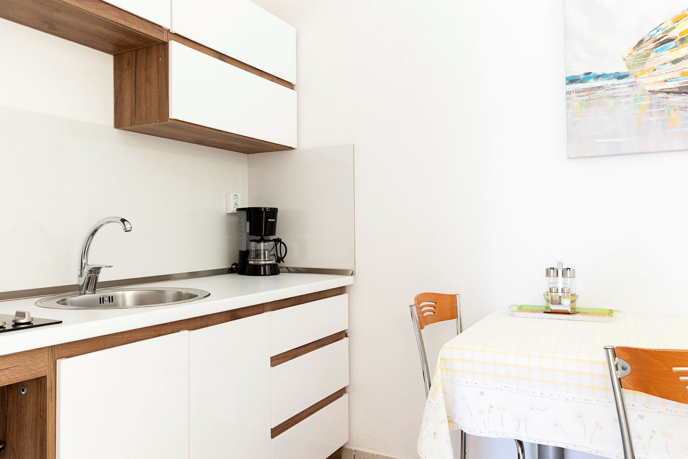 Ett kök med vit inredning, en diskbänk, en kaffebryggare och ett matbord med två stolar. (AI-genererad text)