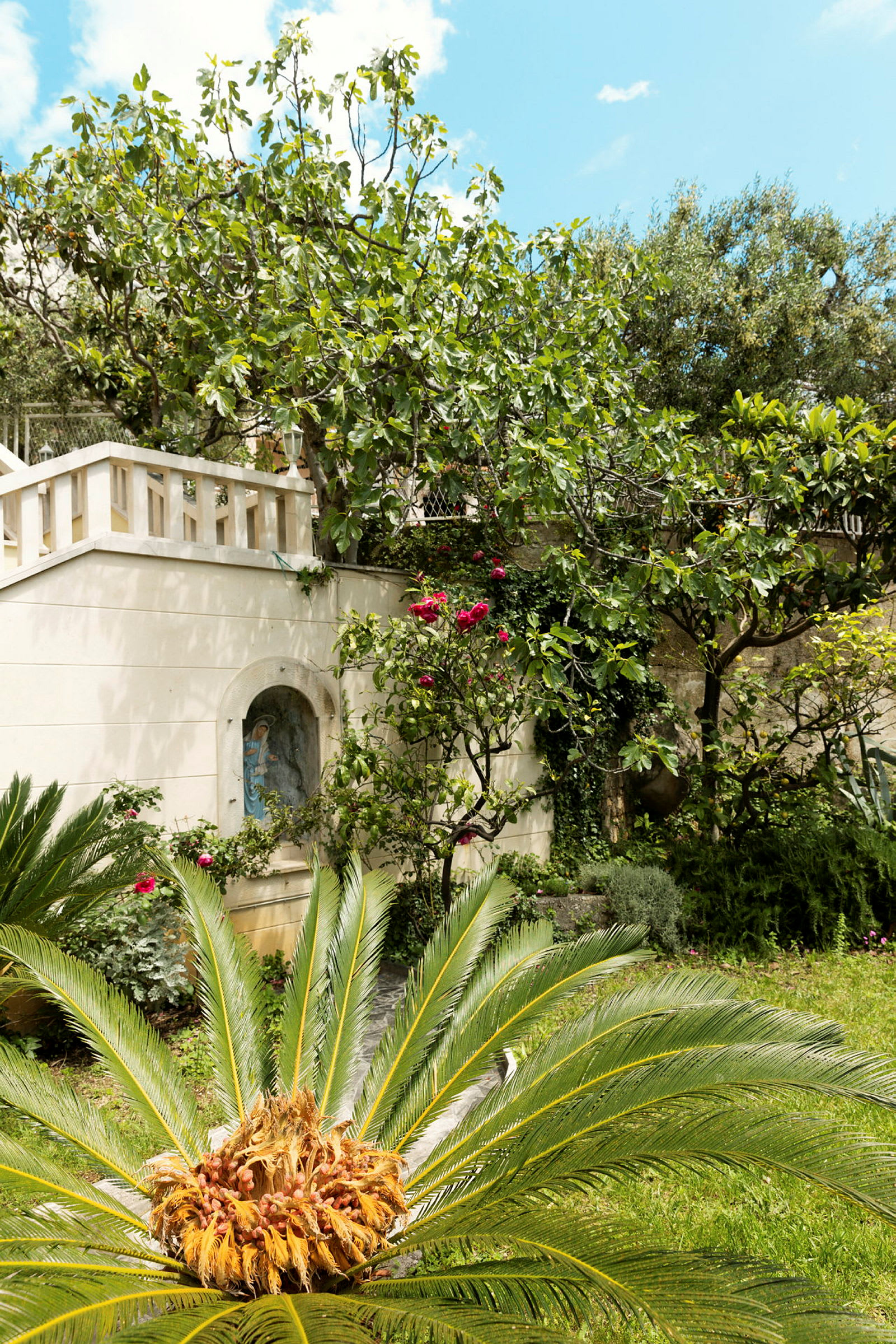En grönskande trädgård med palm och blommande växter vid en byggnad med en skulpturdetalj. (AI-genererad text)