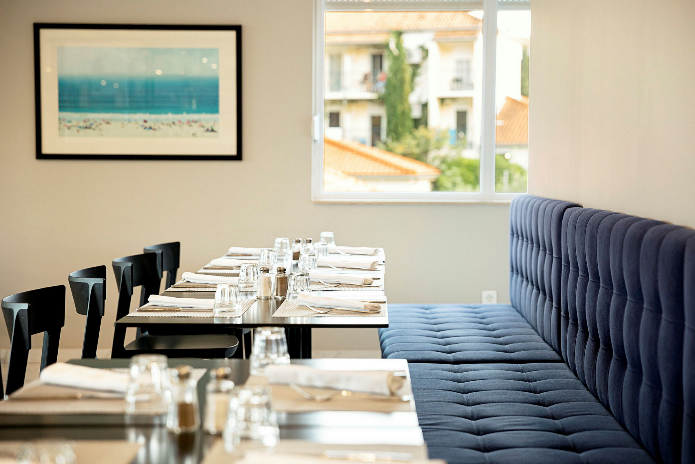 En restaurangmiljö med dukade bord med glas, bestick och servetter. En blå vadderad soffa och fönster som visar byggnader utanför. En tavla med strandmotiv på väggen. (AI-genererad text)