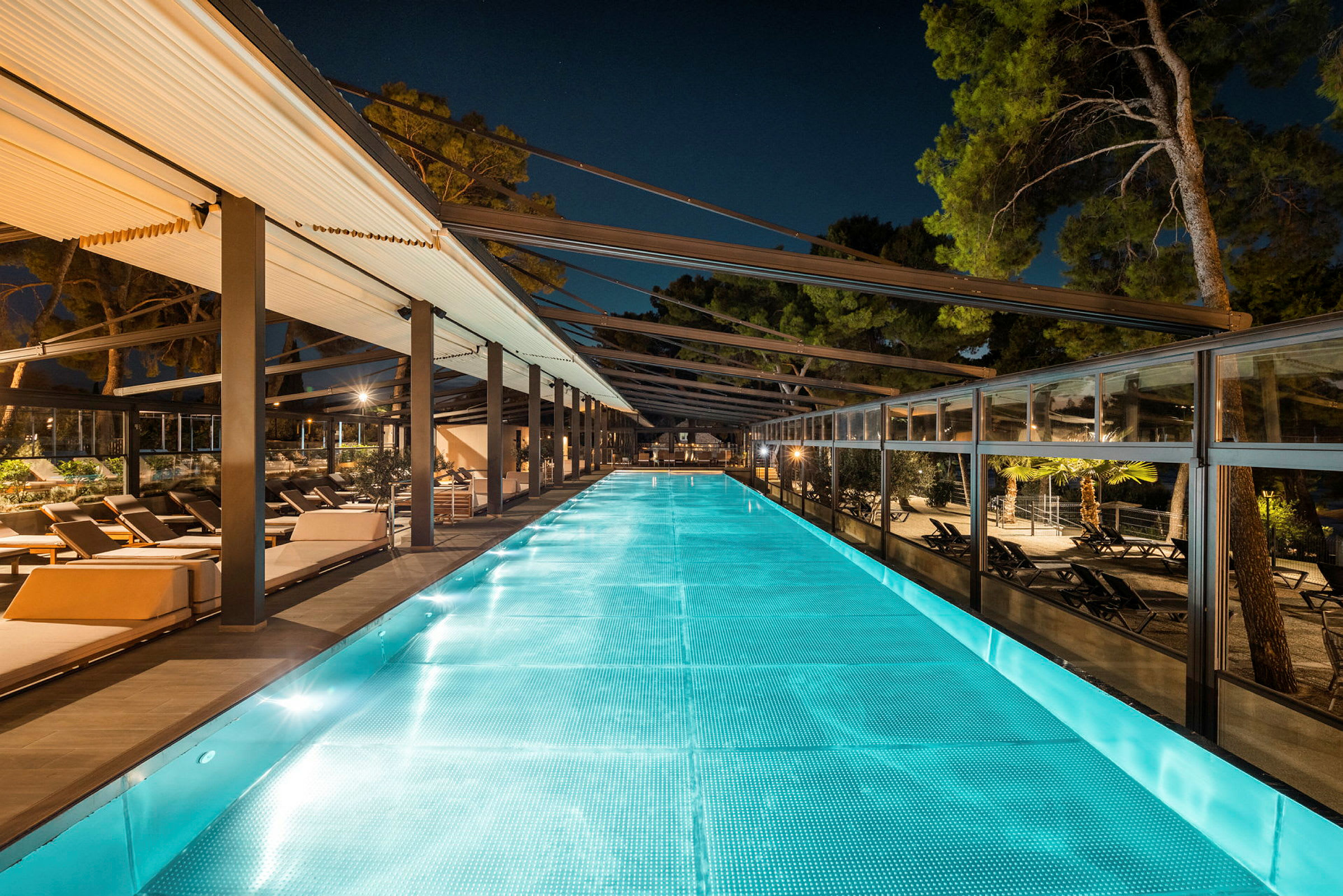 Upplyst pool med sittplatser och omgivande träd på kvällen. (AI-genererad text)