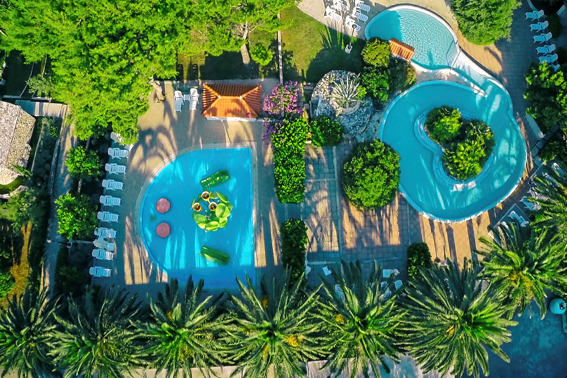 Vy ovanifrån av två swimmingpooler omgivna av stolar, vegetation och gångvägar. (AI-genererad text)