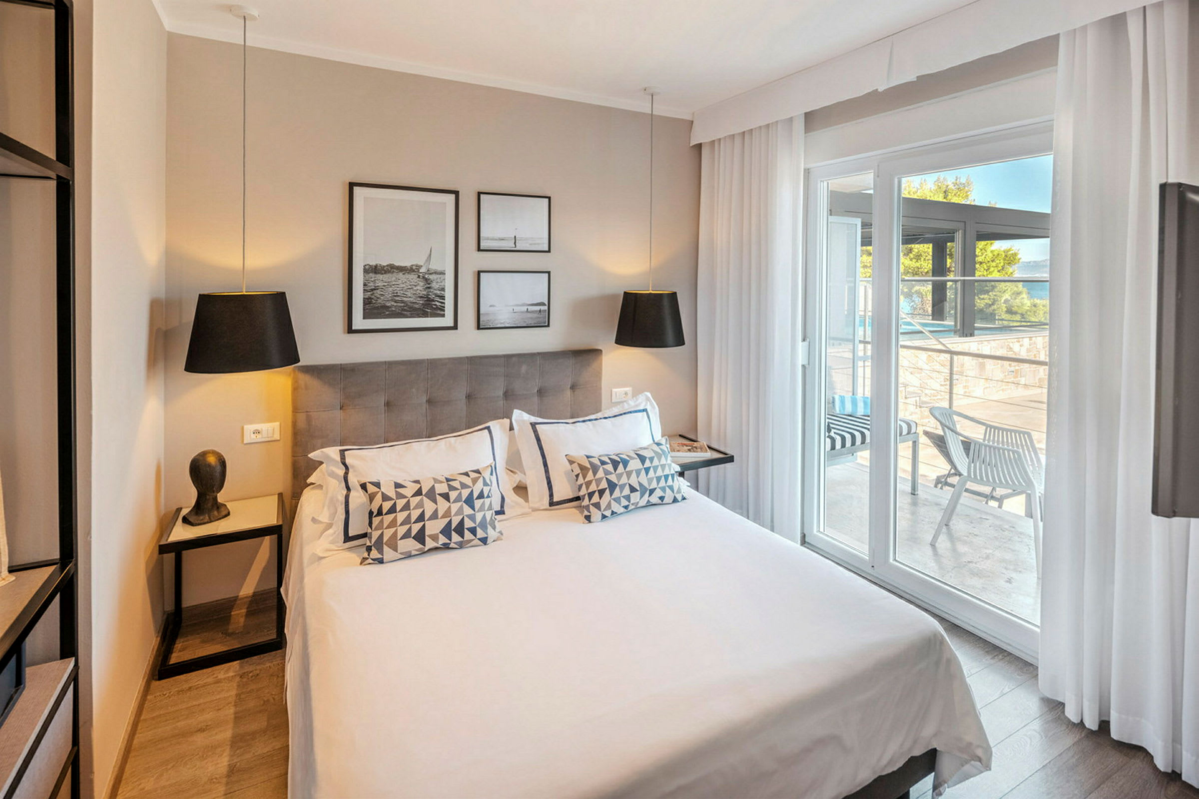 Modernt hotellrum med dubbelsäng, dekorerat med kuddar och konst på väggen. Glasdörrar leder ut till en möblerad terrass. (AI-genererad text)