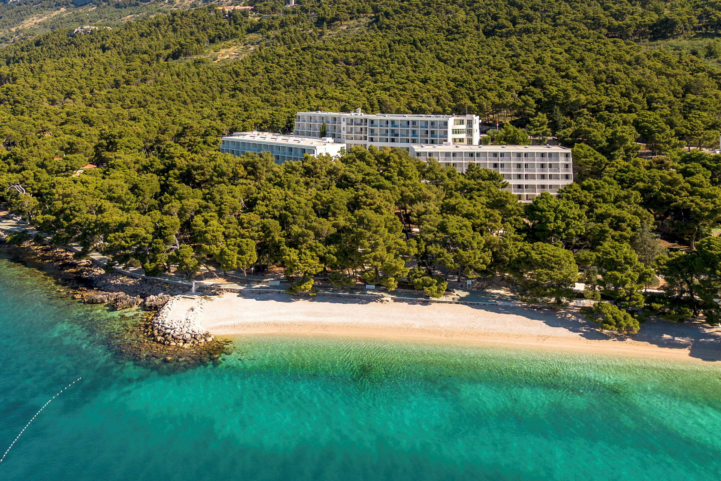 Hotellbyggnader omgivna av grönska nära en strand med klart turkost vatten. (AI-genererad text)