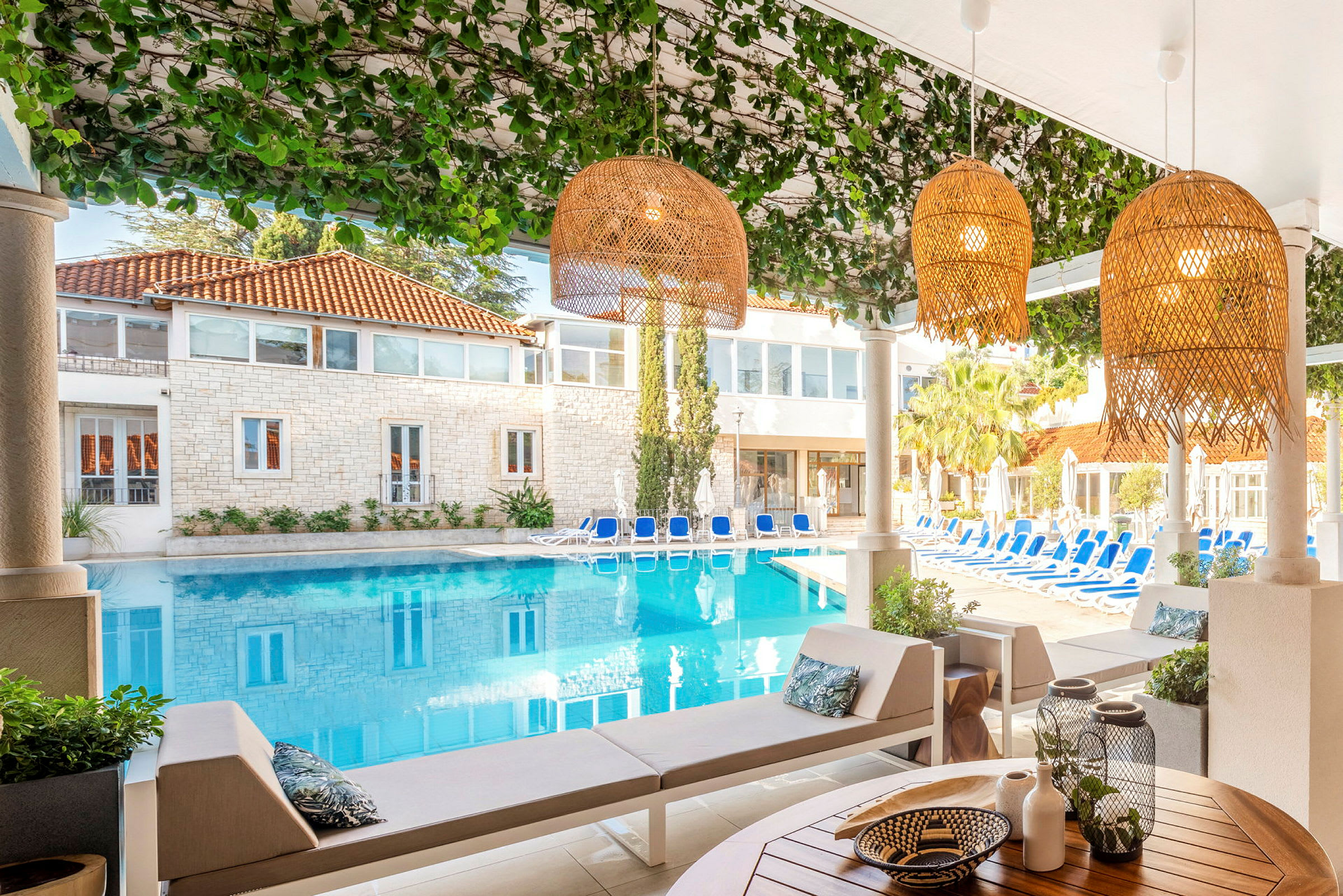 Utsikt över en pool omgiven av solsängar och ett hus med stenfasad, fotograferad från ett skuggat område med sittplatser och dekorationer. (AI-genererad text)