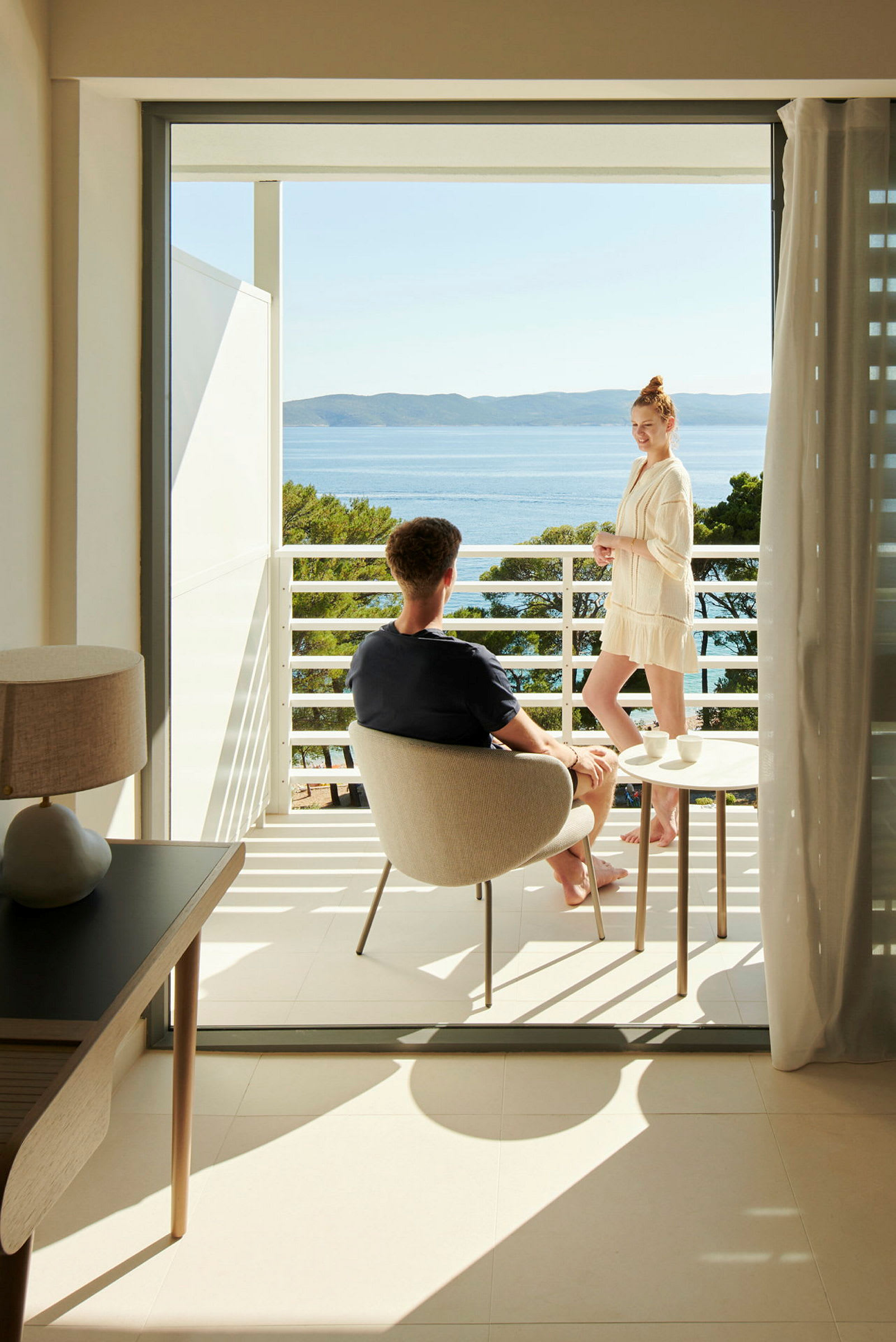 En hotellbalkong med utsikt över havet och omgivande grönska. En person sitter och en annan står bredvid ett bord. (AI-genererad text)