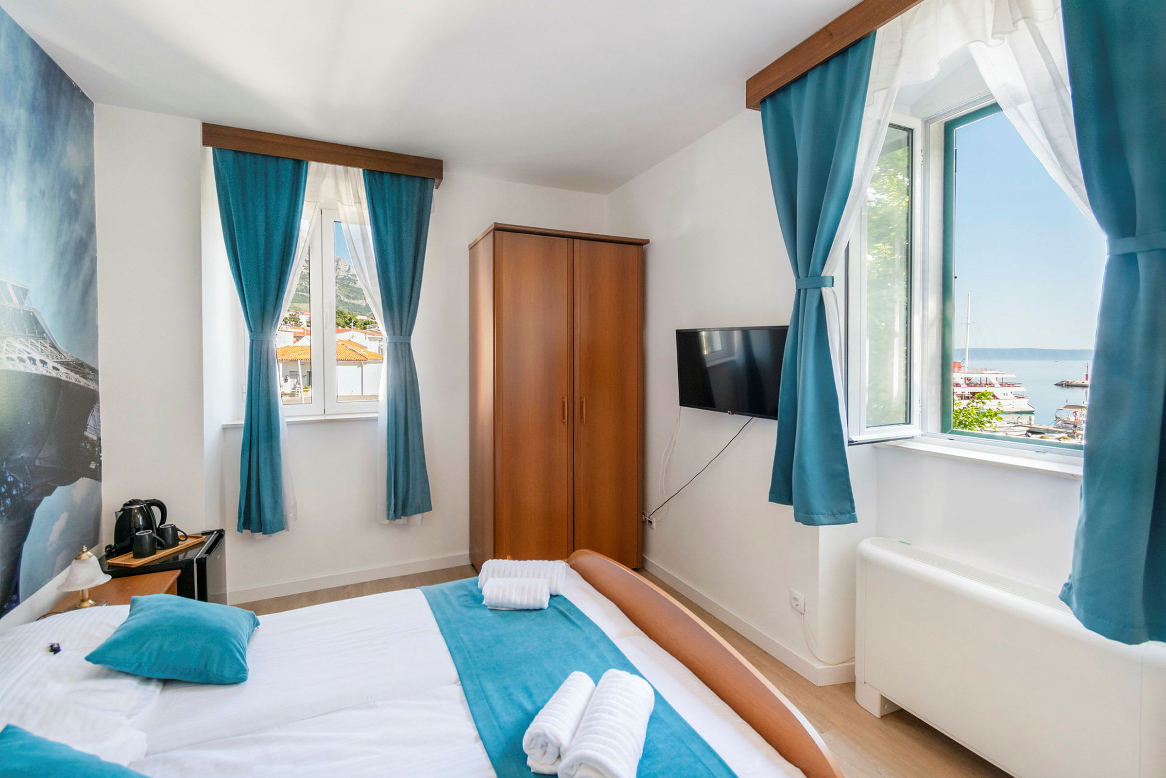 Hotellrum med dubbelsäng, blåa gardiner och utsikt över en hamn utanför fönstret. (AI-genererad text)