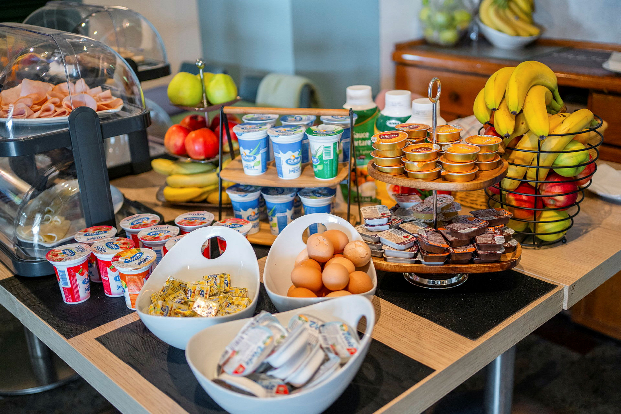 Bilden visar ett frukostbord med olika matvaror som ägg, yoghurt, frukt, pålägg och smörförpackningar. (AI-genererad text)