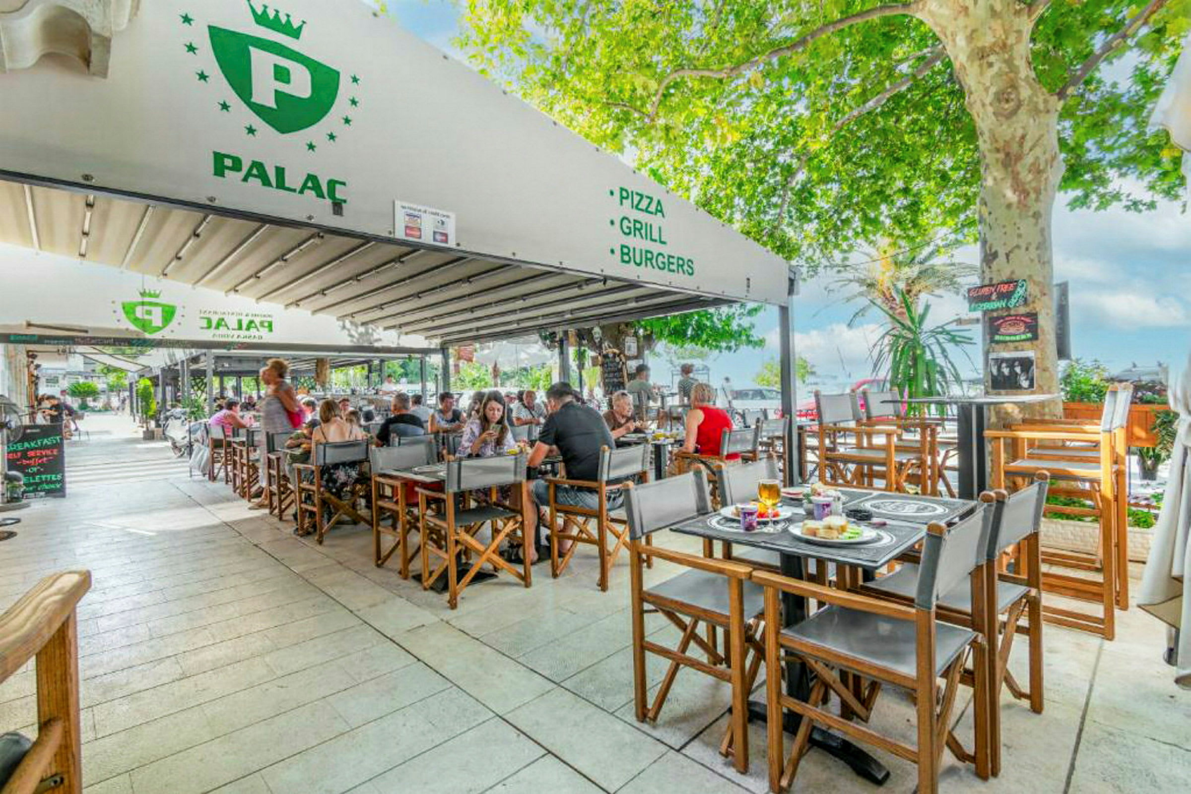 Uteservering vid restaurang med gäster vid bord, omgivet av grönska. (AI-genererad text)