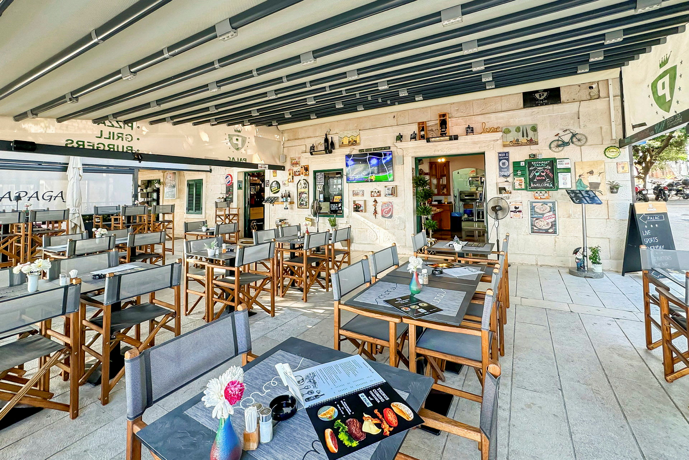 Restaurang med uteservering, bord och stolar, samt dekorationer och en TV på väggen. (AI-genererad text)