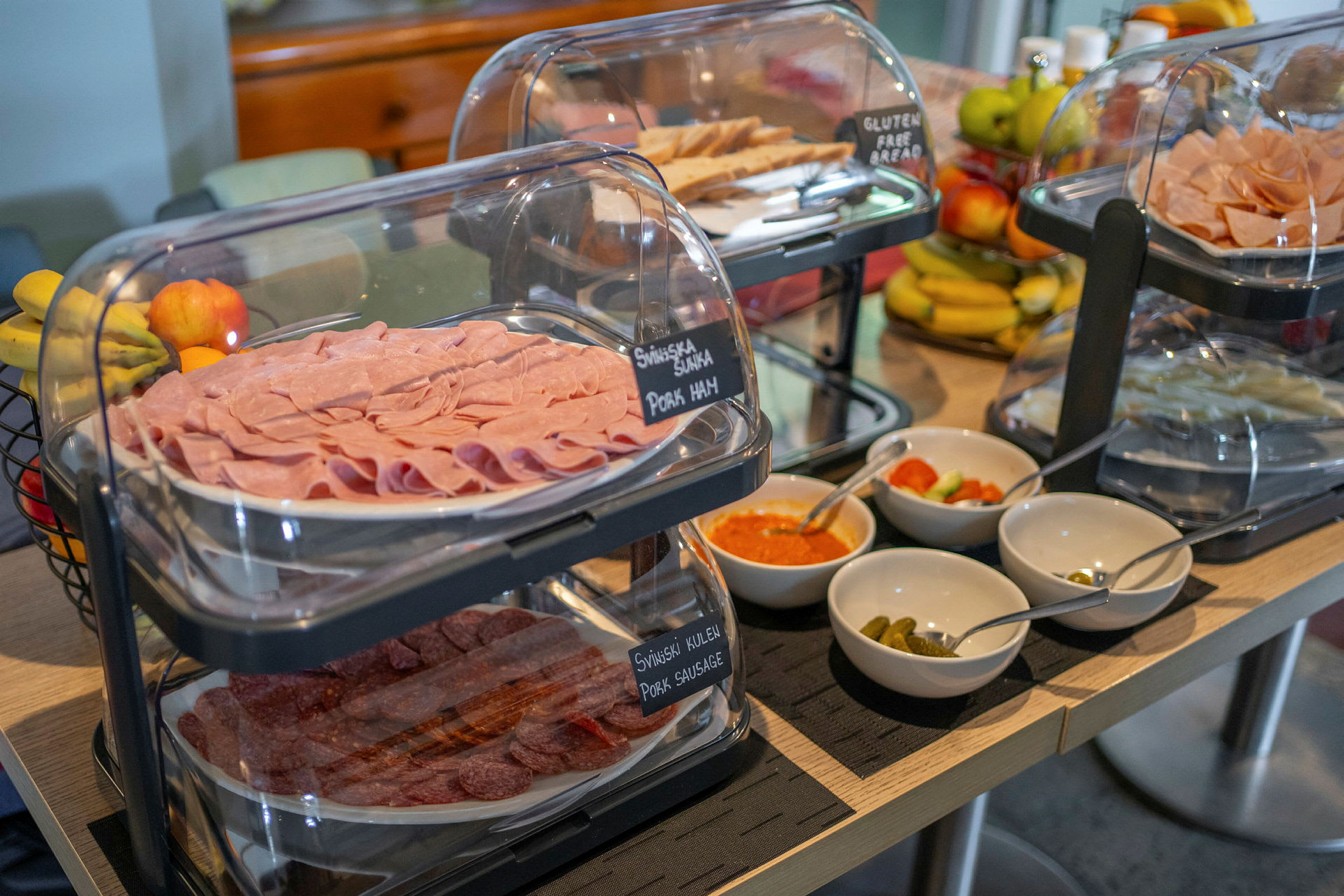 Buffébord med skivor av skinka och korv, ett urval av röror och grönsaker samt frukt i bakgrunden. (AI-genererad text)