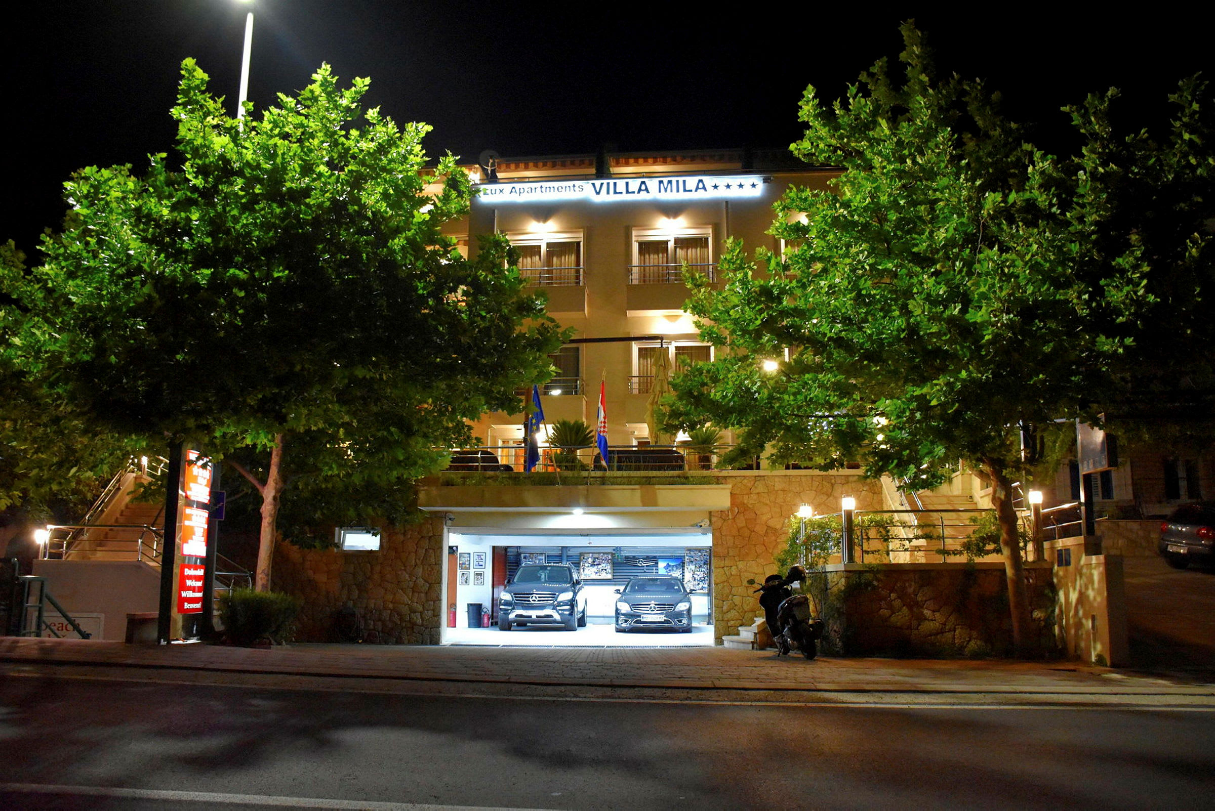 Hotellbyggnad med upplyst entré och parkeringsgarage, omgiven av träd, på natten. (AI-genererad text)
