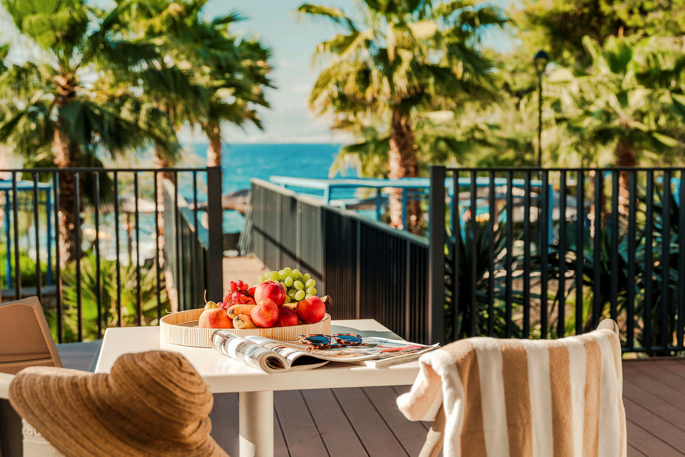 Ett dukat bord med fruktfat, en tidning och ett par solglasögon, med utsikt mot en terrass med palmer och havet i bakgrunden. (AI-genererad text)