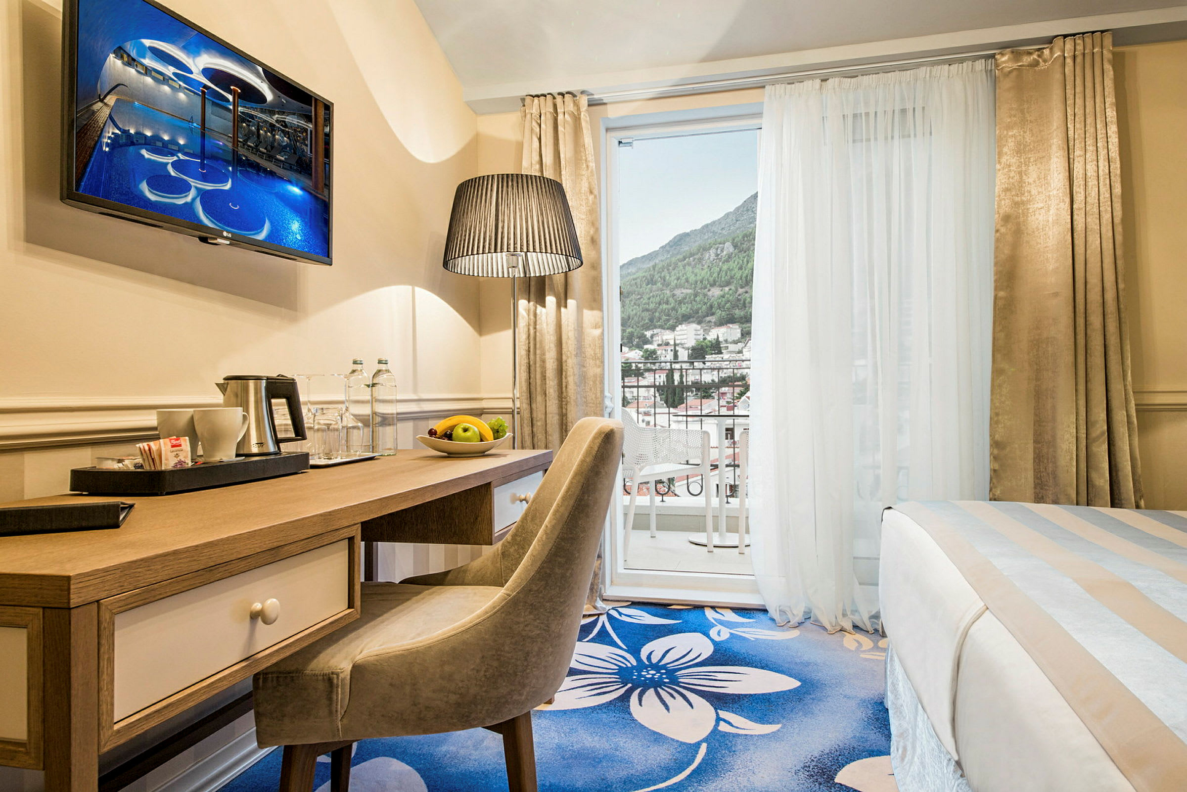 Ett hotellrum med ett skrivbord, stol och en säng, samt en öppen balkongdörr mot en utsikt över grönklädda berg och byggnader. (AI-genererad text)