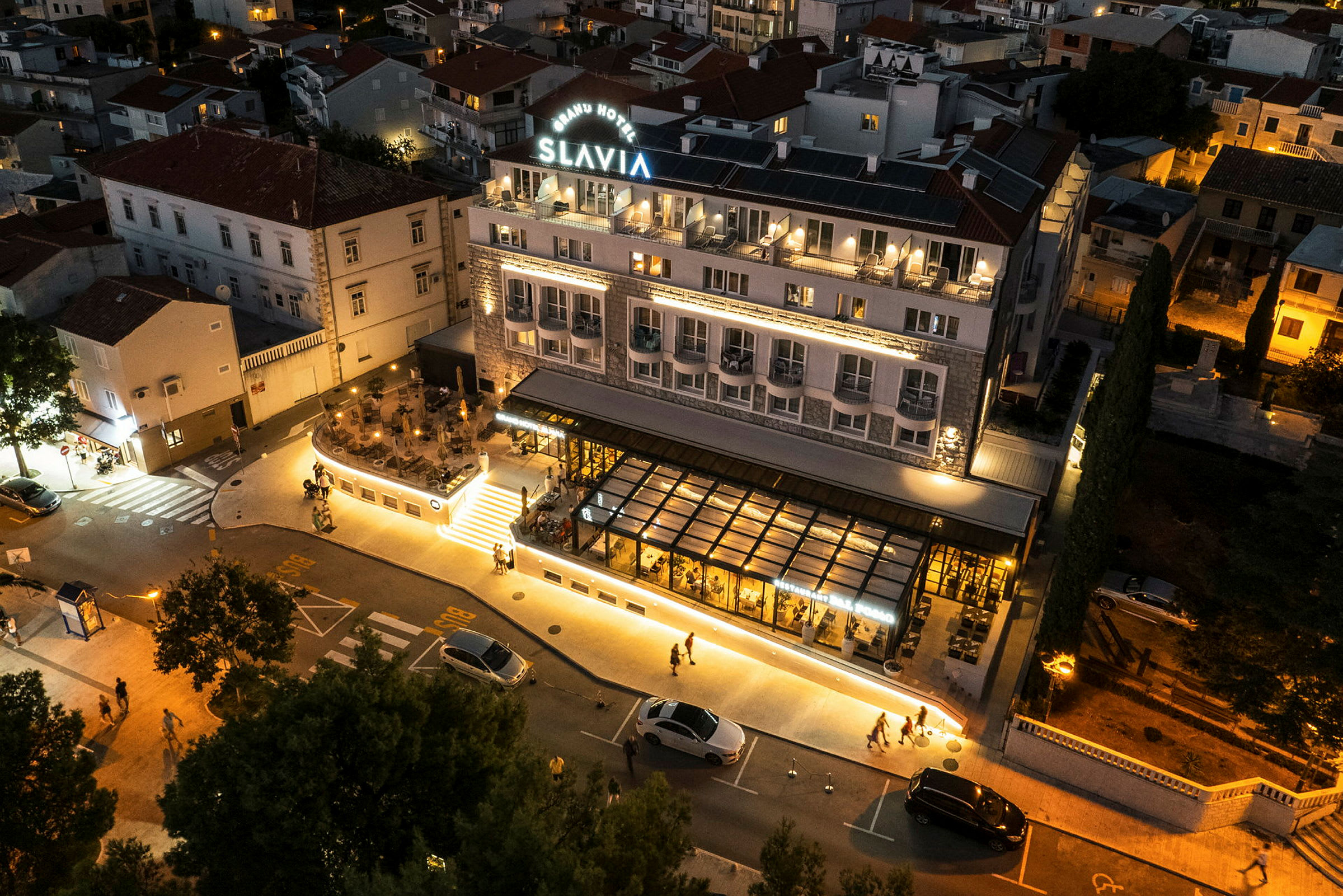 Ett hotell med namnet "Slavia" syns upplyst på kvällen. Byggnaden har balkonger och en uteservering. En väg och flera parkerade bilar ses i förgrunden. (AI-genererad text)