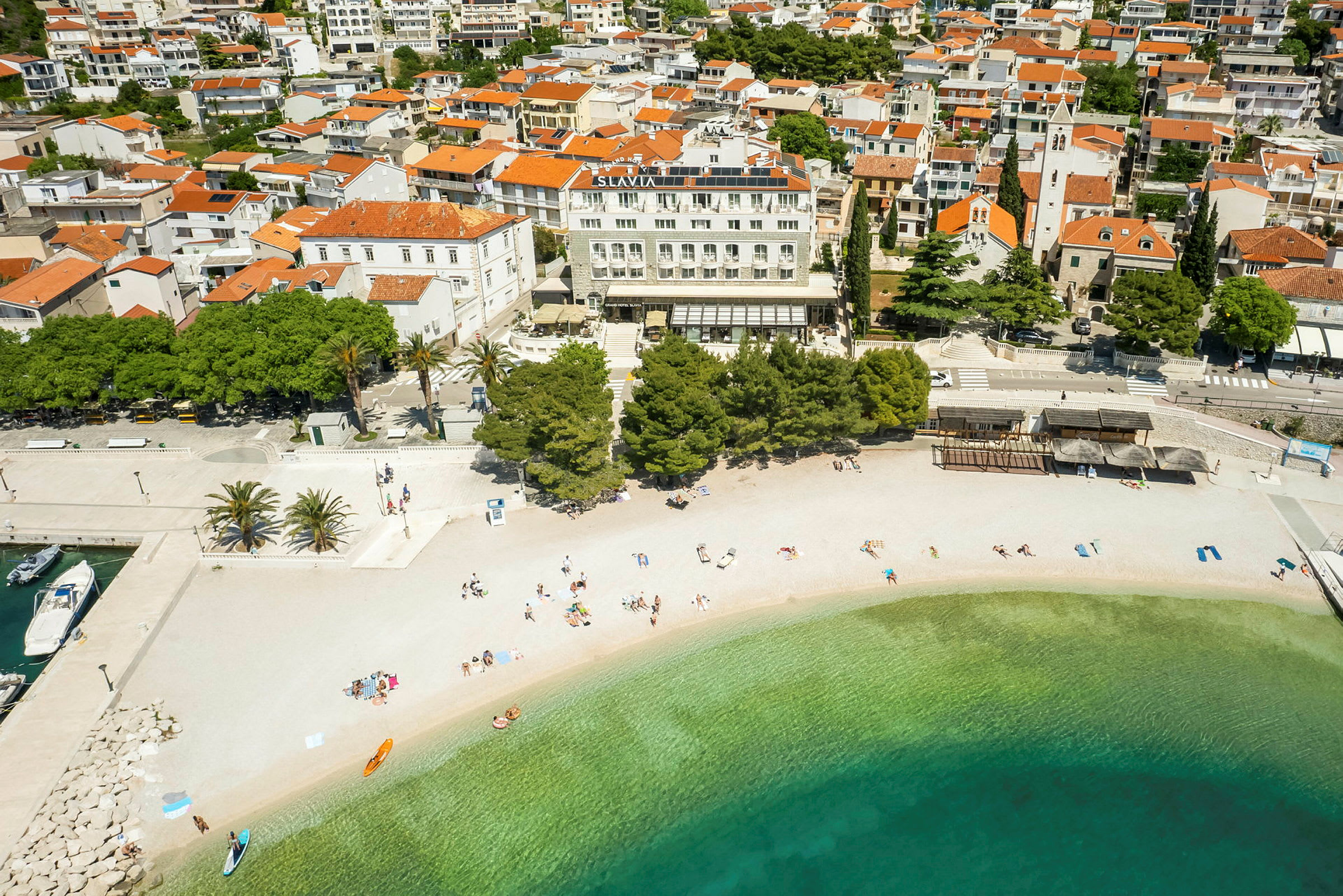 Flygbild av en strand med klart vatten framför en stad med röda tegeltak på hus och byggnader. (AI-genererad text)