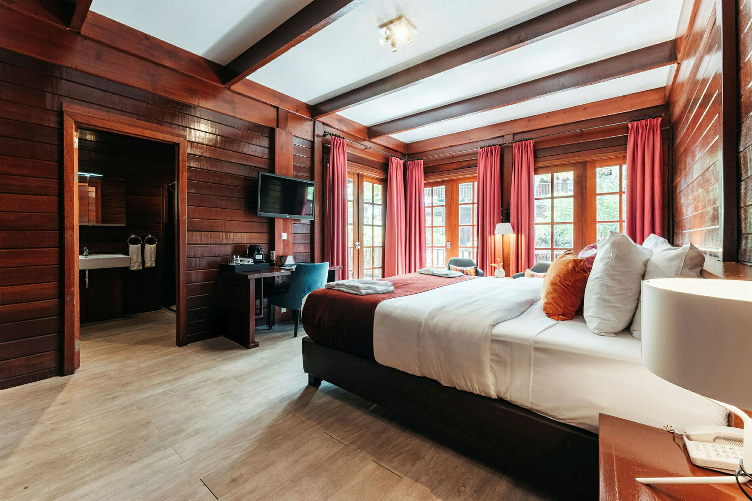 Hotellrum med träinredning, en bred säng med vita och färgglada kuddar, röda gardiner och fönster med utsikt. (AI-genererad text)