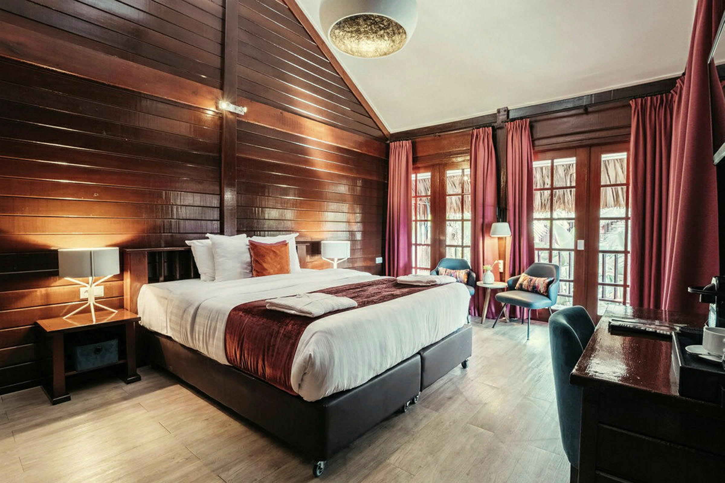 Ett mysigt hotellrum med träväggar och stora fönster täckta av röda gardiner. Rummet har en dubbelsäng med vita sängkläder och dekorativa kuddar. (AI-genererad text)