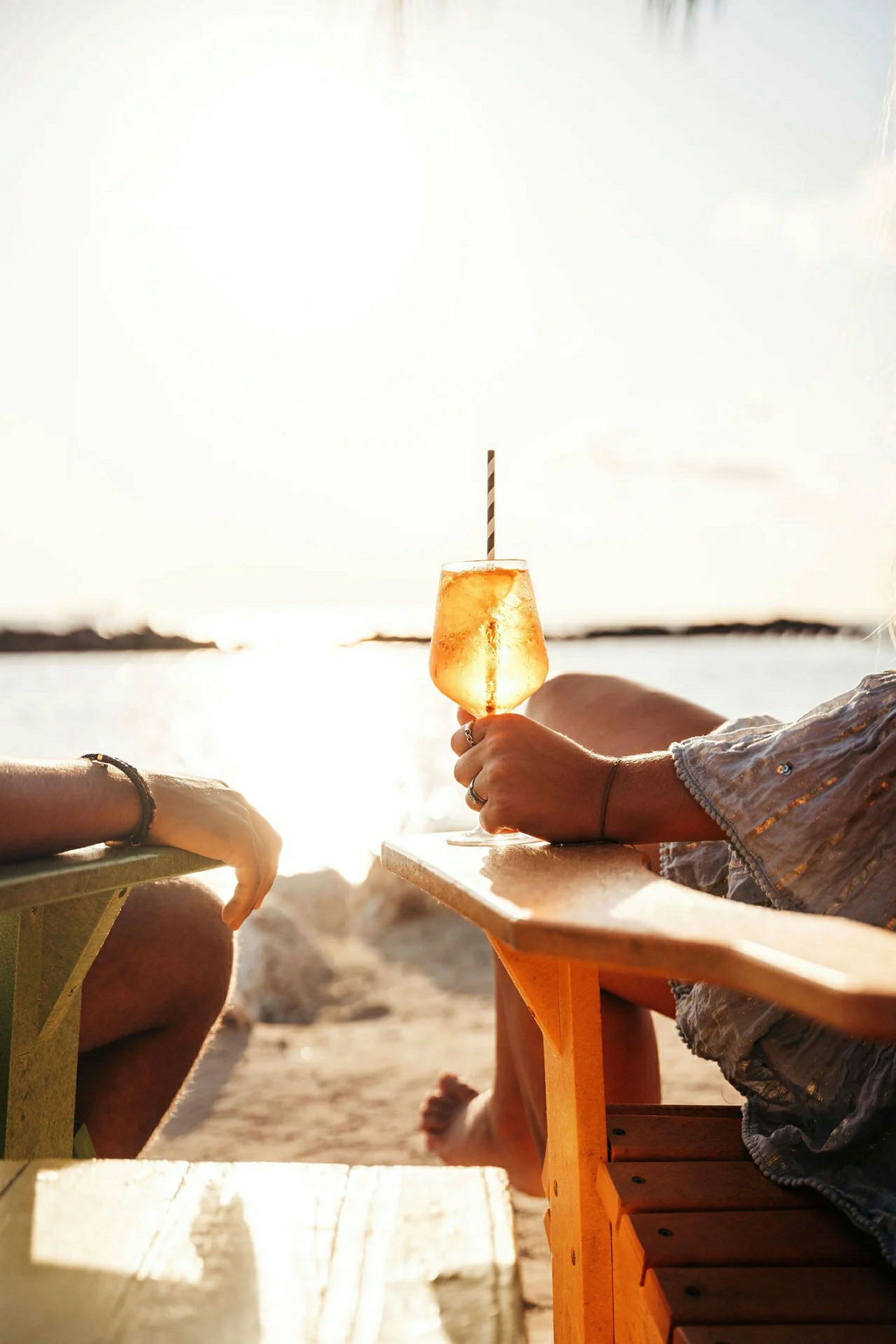 Två personer sitter i stolar på en strand med utsikt över havet och solnedgången. I förgrunden hålls ett glas med en drink. (AI-genererad text)