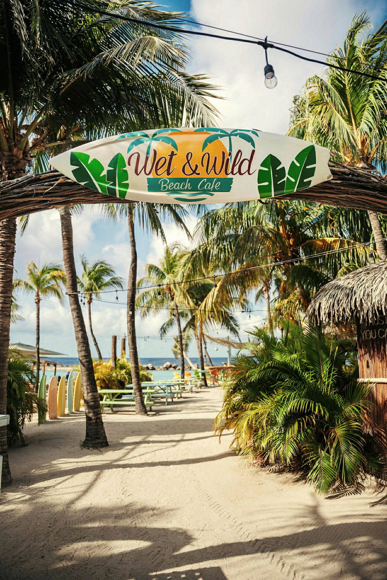 Skylt med texten "Wet & Wild Beach Café" hänger på en båge i en utomhusmiljö med palmer, bord och hav i bakgrunden. (AI-genererad text)