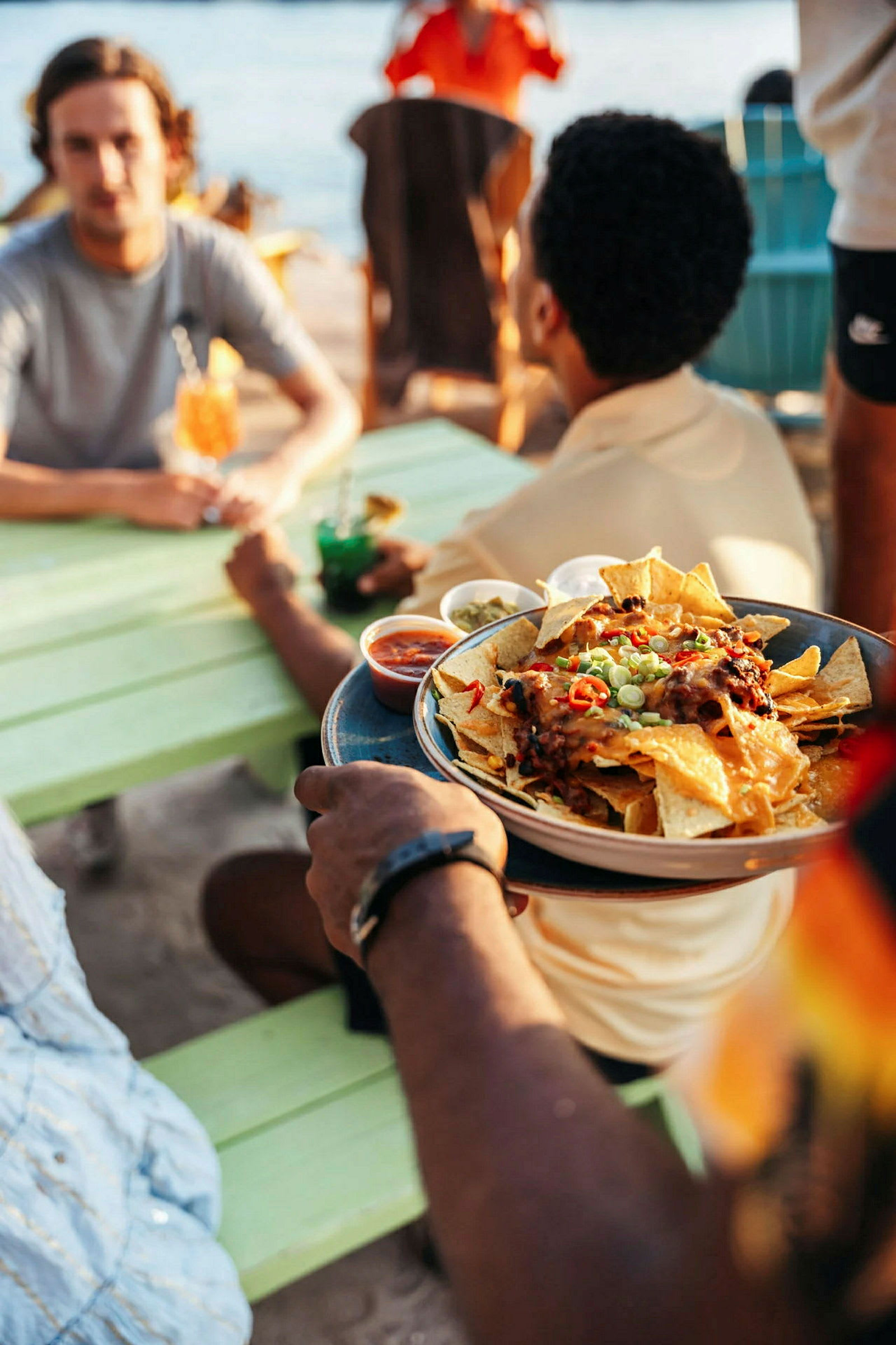 En person håller en tallrik med nachos, olika dipsåser syns runt tallriken. Scenen är utomhus vid ett bord nära vatten. (AI-genererad text)