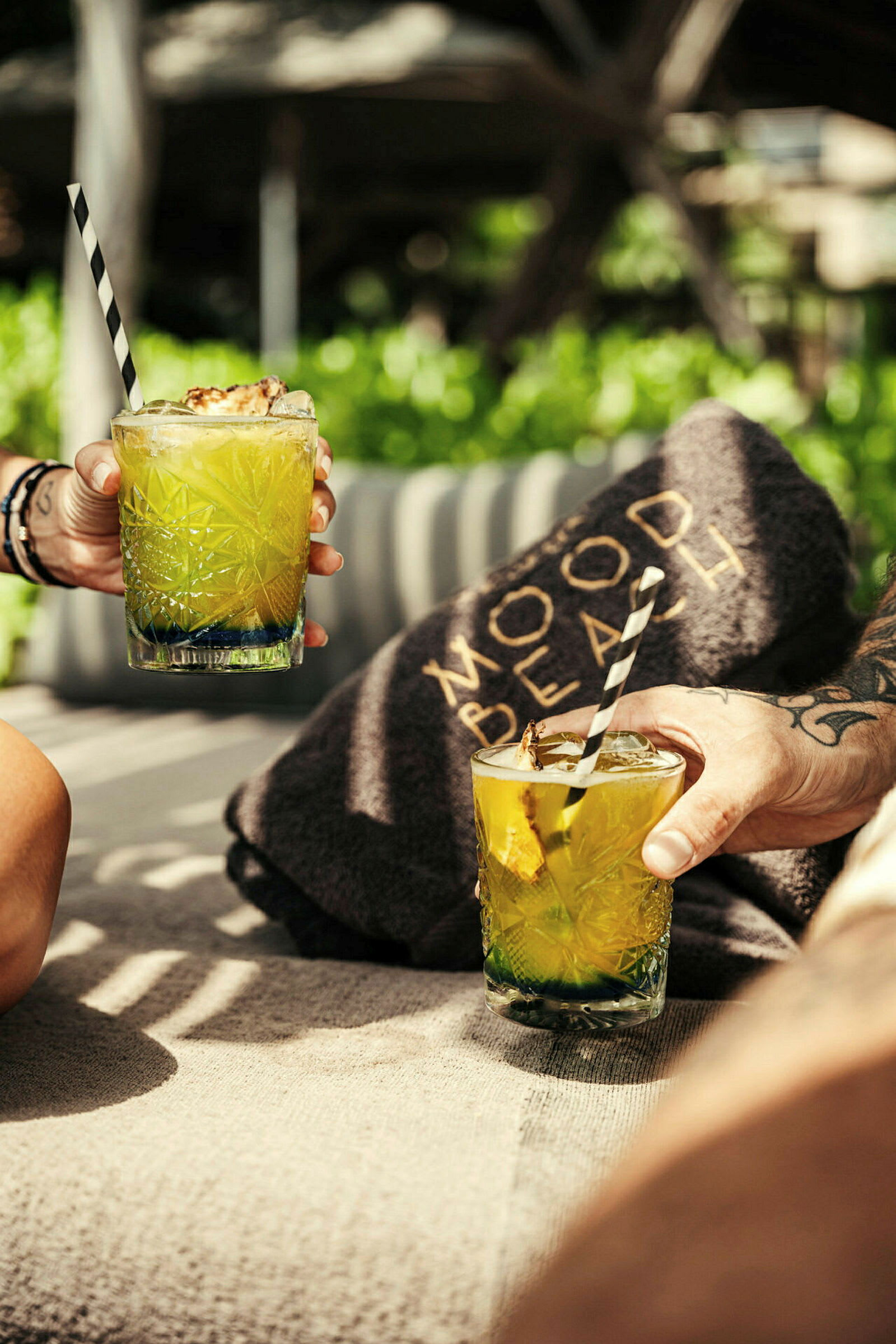 Två händer håller upp glas med gröna drinkar och svartvita sugrör på solstolar med en filt märkt 'Mood Beach' i bakgrunden. (AI-genererad text)