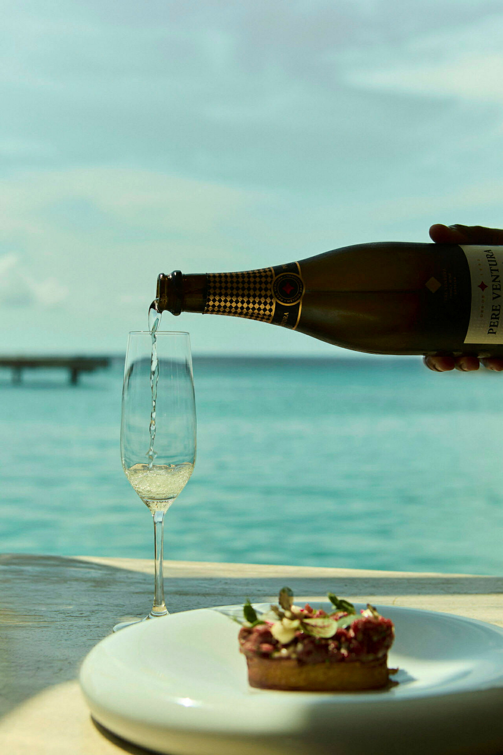Ett glas fylls med champagne bredvid en tallrik med en liten dessert, med havsutsikt i bakgrunden. (AI-genererad text)