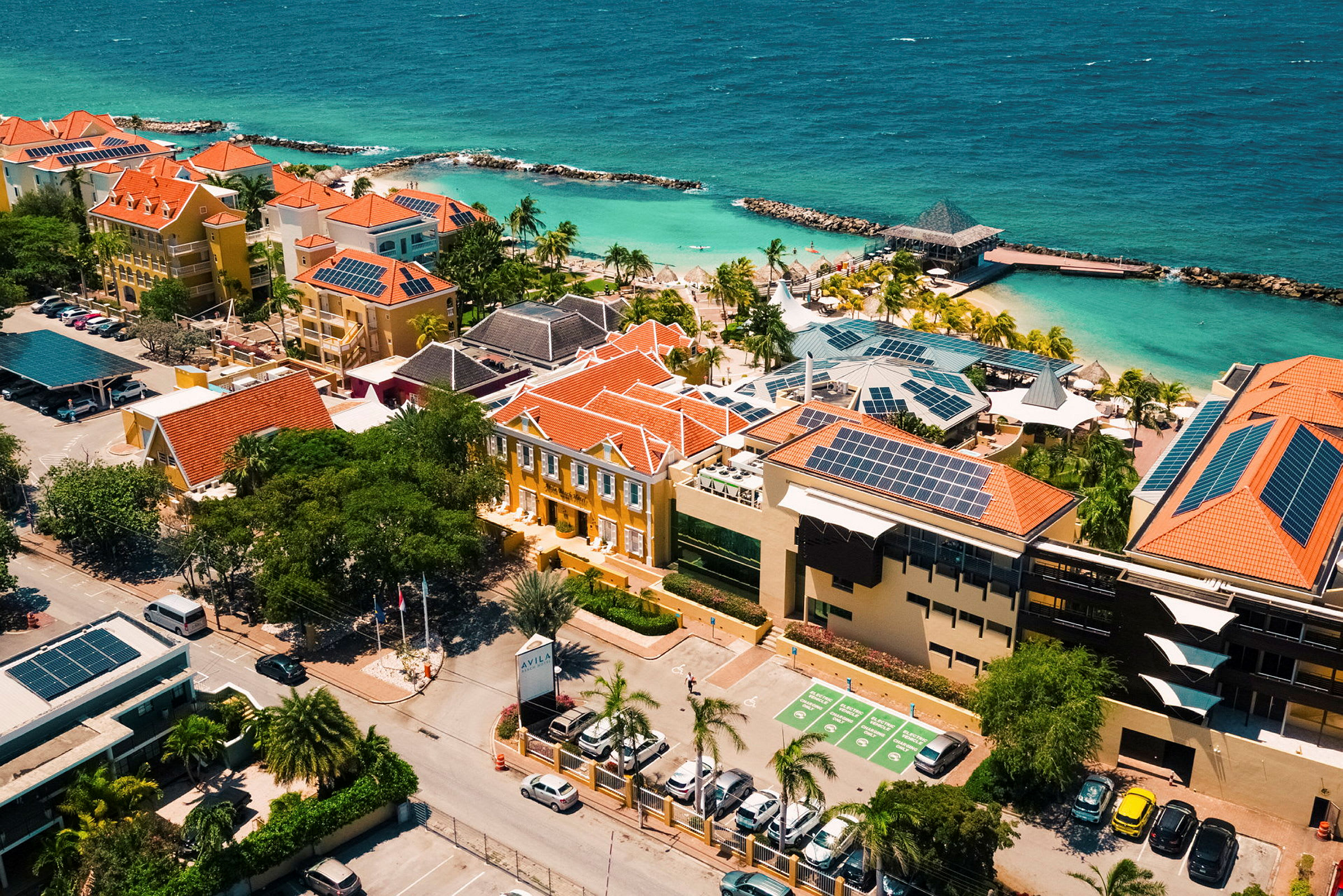 Flygfoto över en semesteranläggning med färgglada byggnader och solpaneler nära en strand med kristallklart vatten. (AI-genererad text)