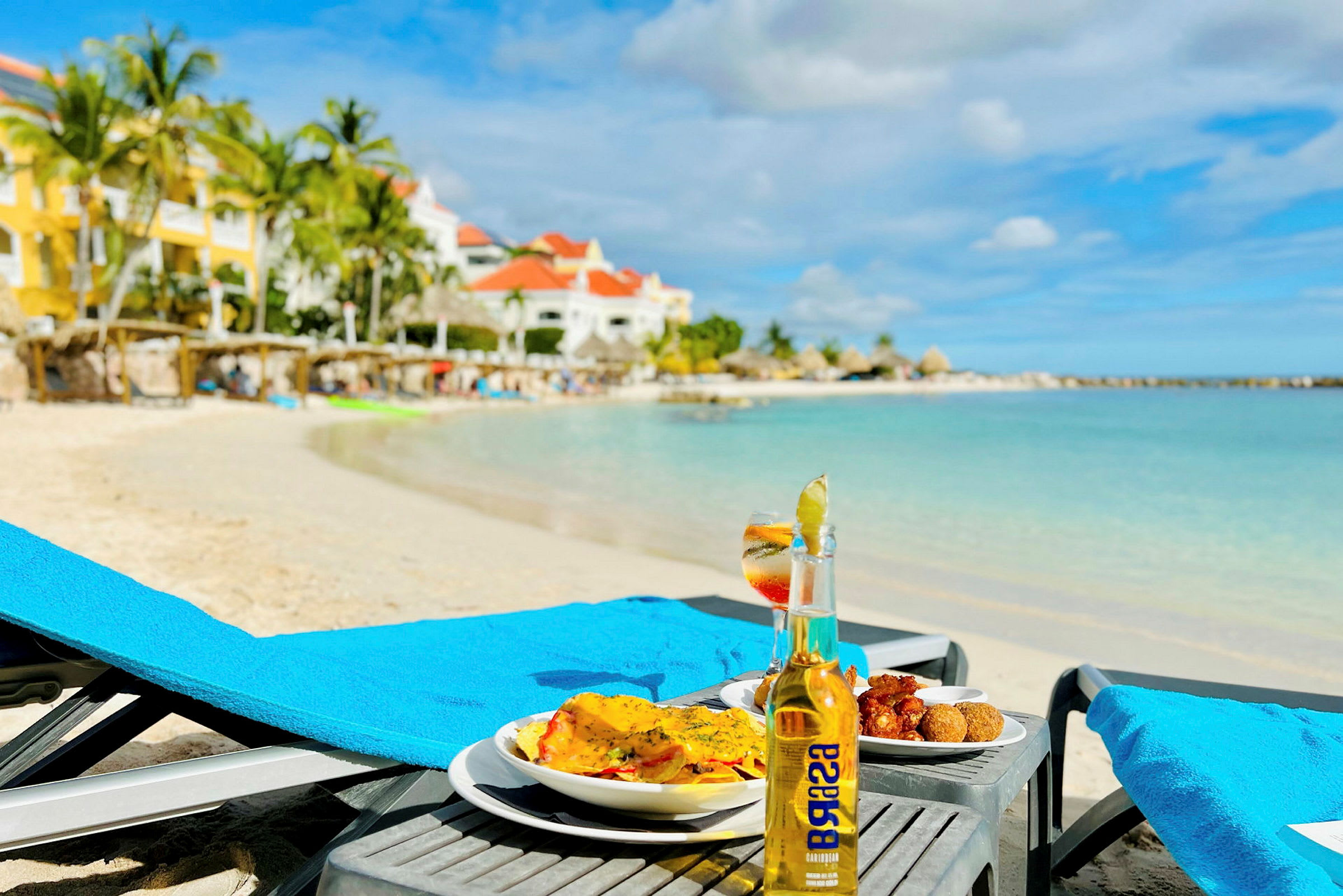 Ett bord med mat och dryck står bredvid en solstol på en sandstrand med turkost vatten. I bakgrunden syns palmer och byggnader. (AI-genererad text)