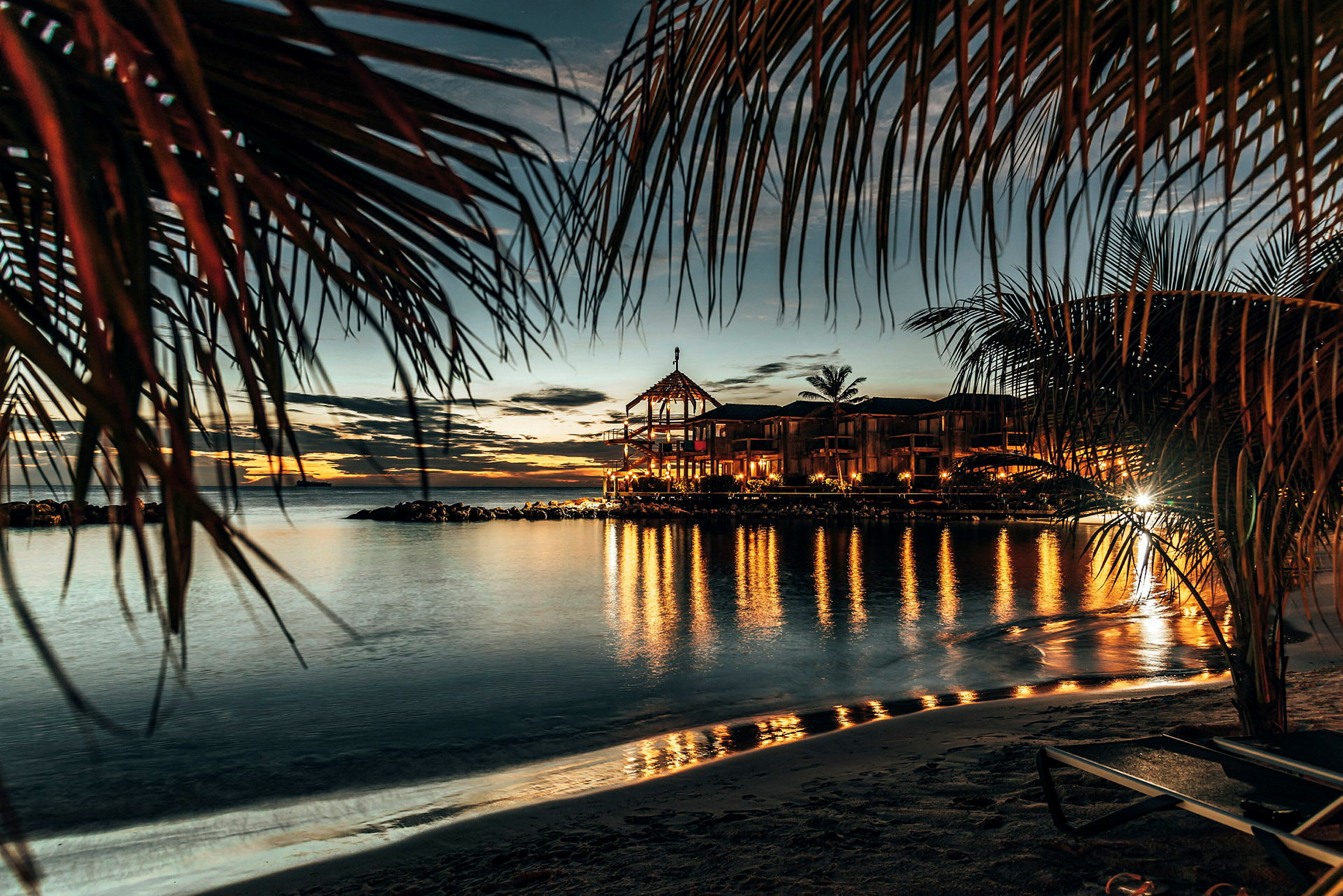 En solnedgång vid havet med palmblad i förgrunden, en byggnad med belysning och dess reflektion i vattnet. (AI-genererad text)
