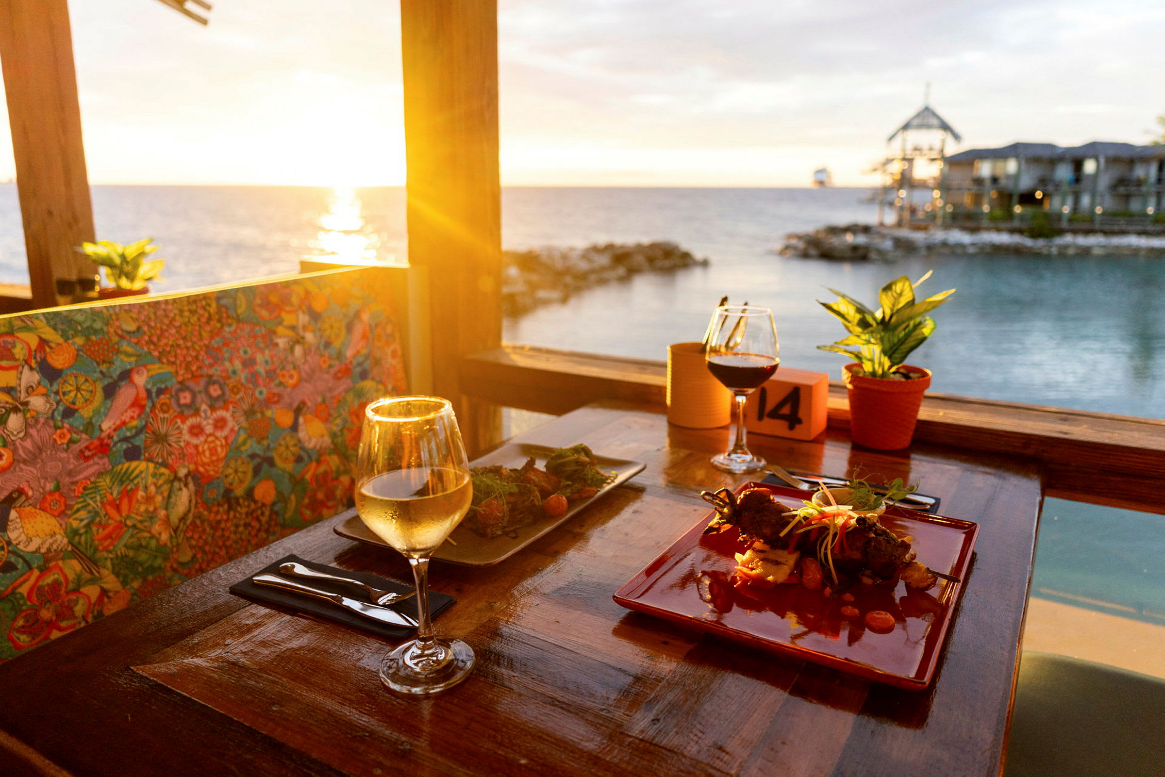 En restaurangstolsrygg med färgglatt mönster, ett välserverat bord med glas vin och mat, utsikt över havet i solnedgången. (AI-genererad text)