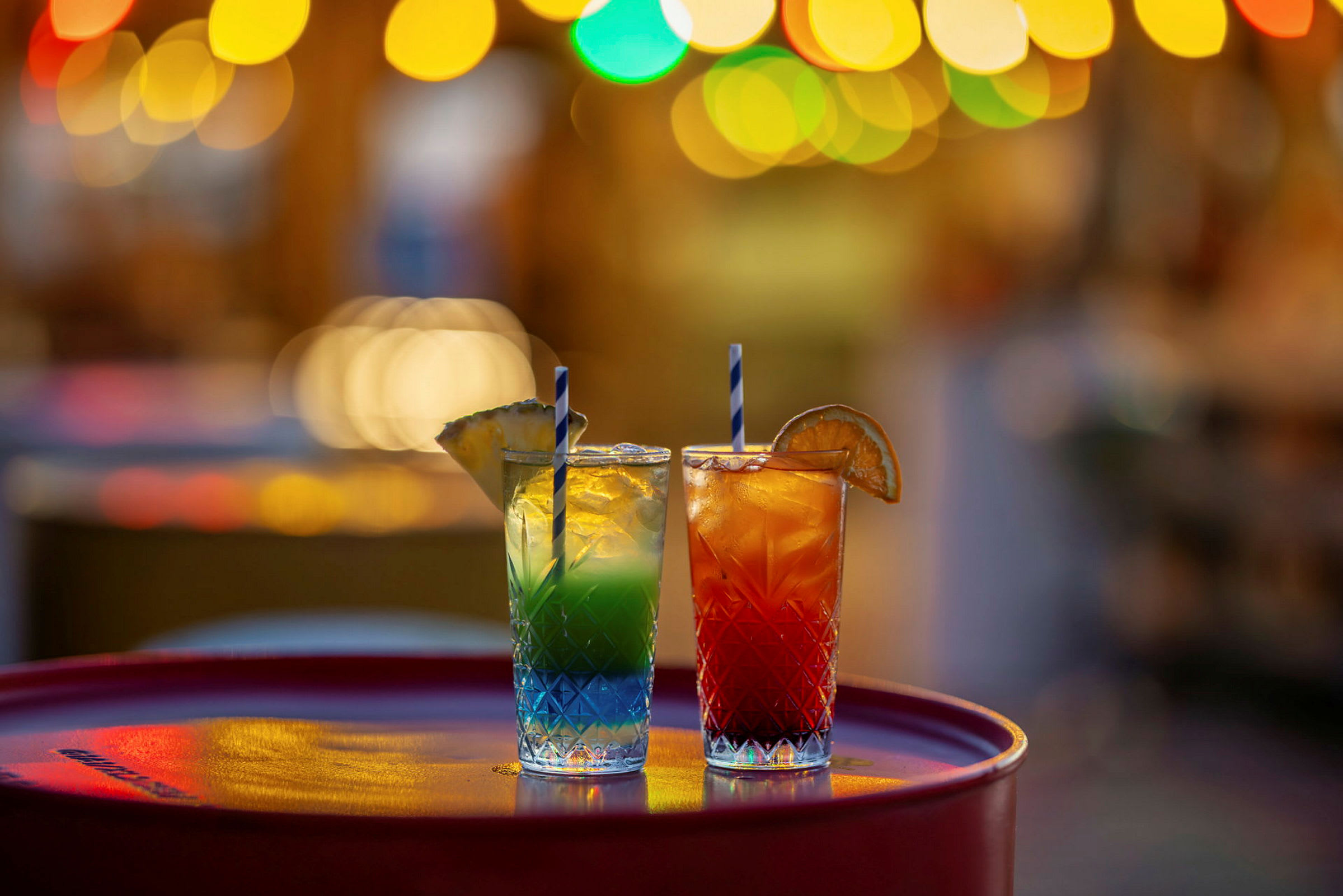 Två glas med färgglada cocktails, dekorerade med frukt, står på ett bord med oskarpa ljus i bakgrunden. (AI-genererad text)