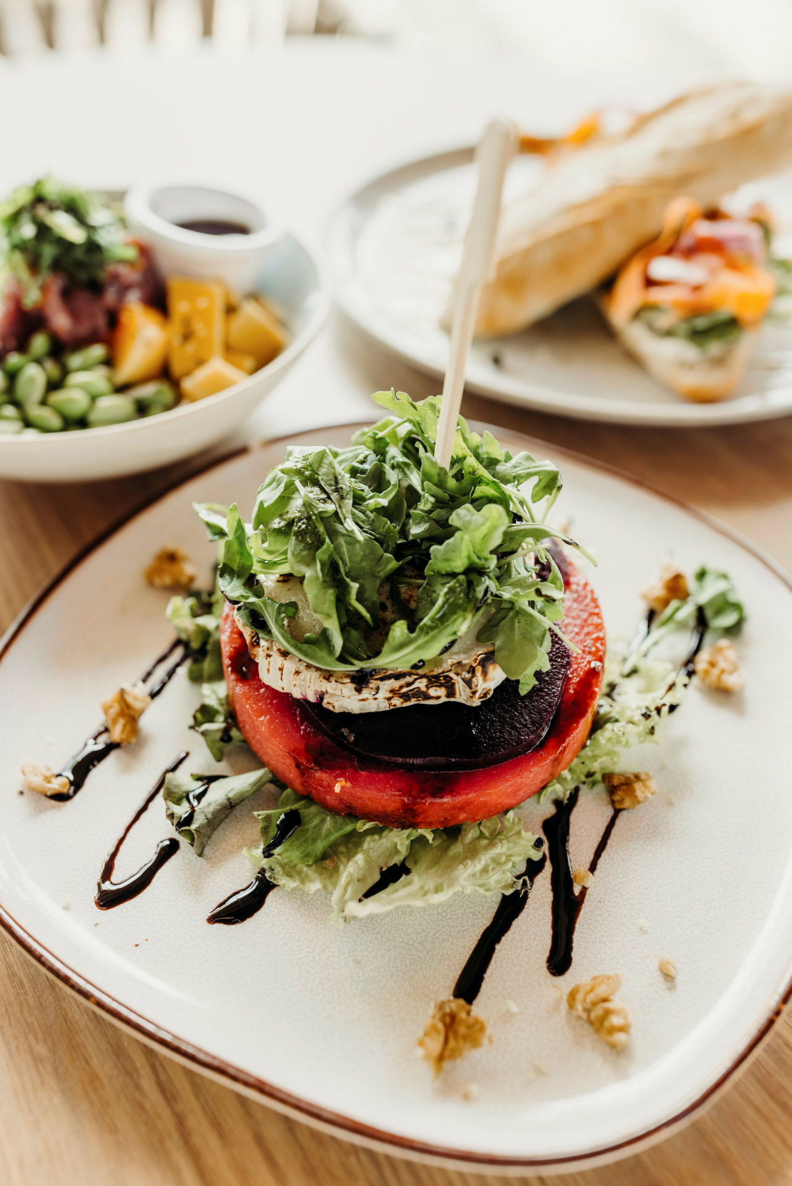 Tallrik med en sallad innehållande grillad grönsak, ost och ruccola, dekorerad med balsamico och nötter. Bakom syns en brödrätt och en skål med färgglada ingredienser. (AI-genererad text)