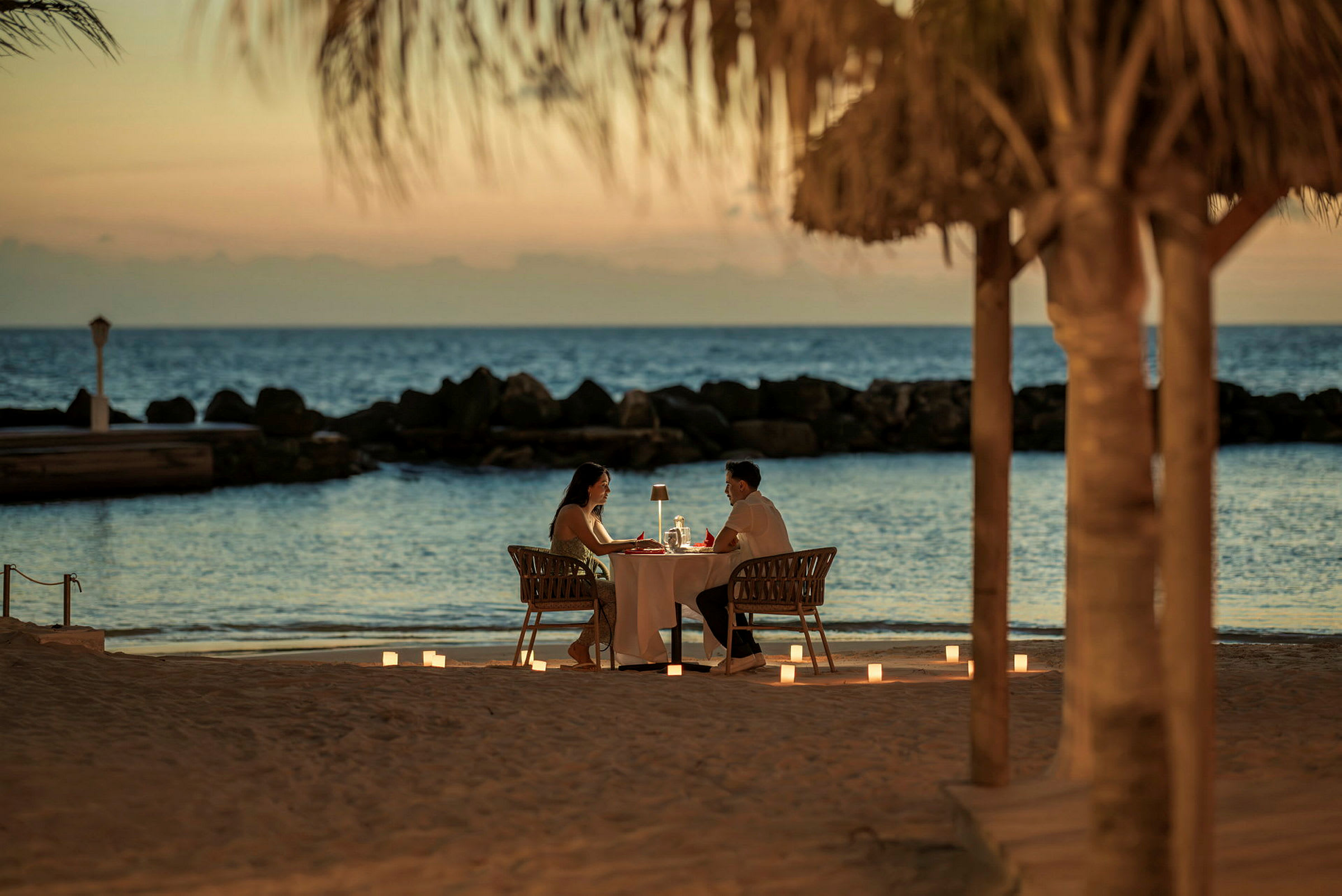 Ett par sitter vid ett dukat bord på stranden med havet i bakgrunden och stearinljus placerade omkring. (AI-genererad text)