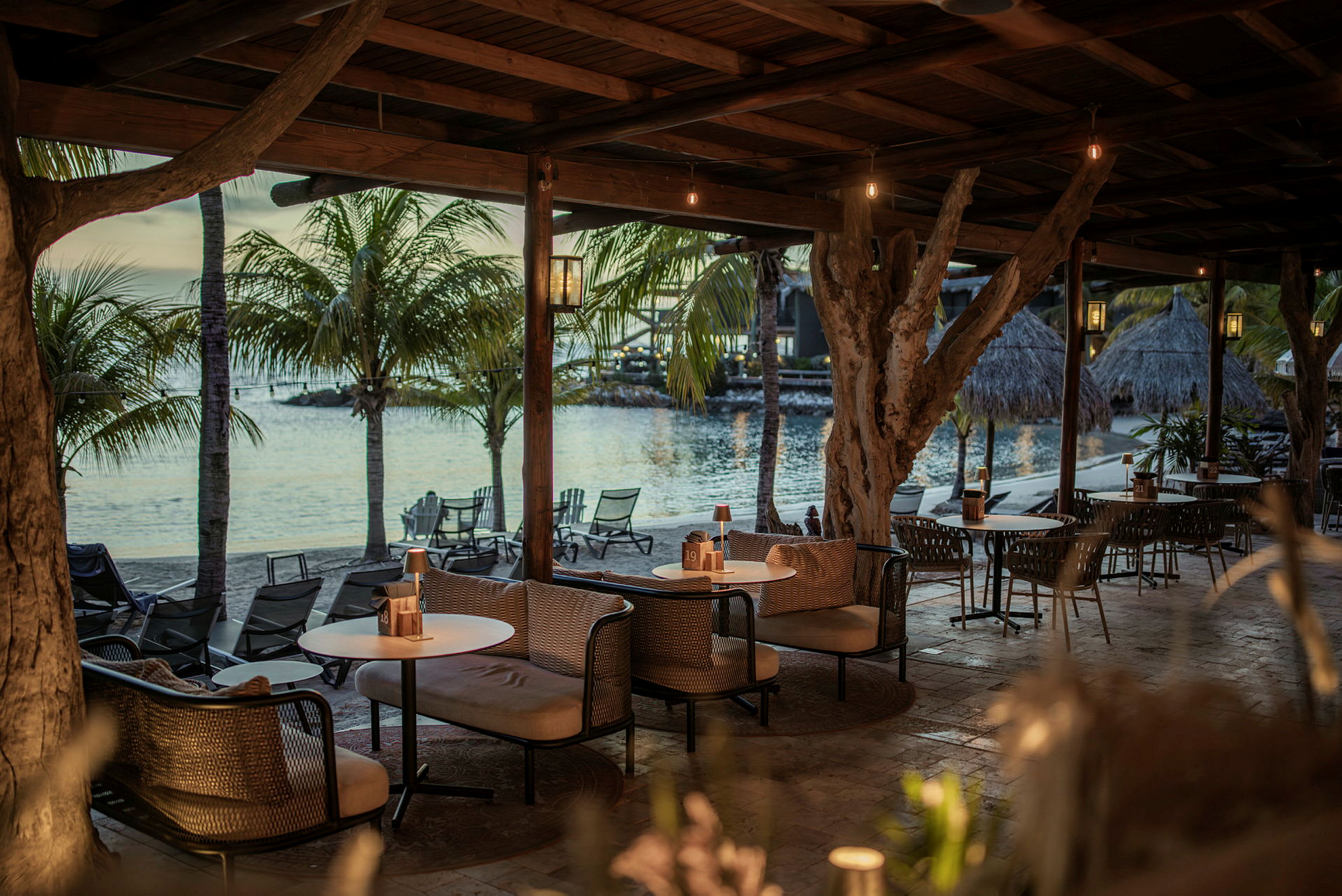 En utomhuslounge under ett träskjul med soffor och bord vid havet, omgiven av palmträd. (AI-genererad text)