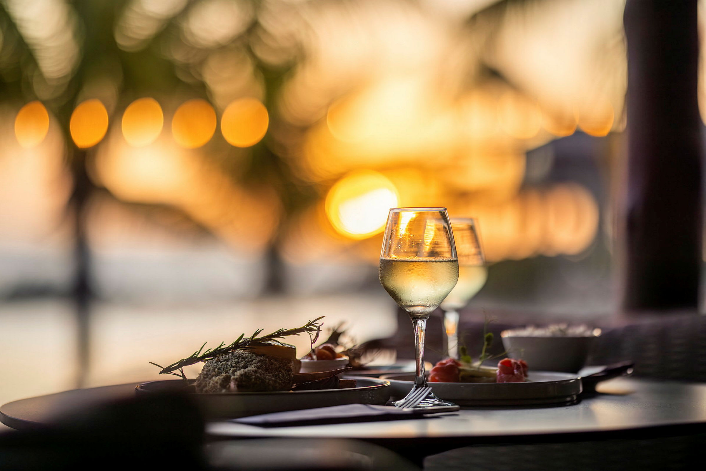 Ett dukat bord med glas vin och tallrikar med mat vid solnedgången, med suddiga palmer i bakgrunden. (AI-genererad text)