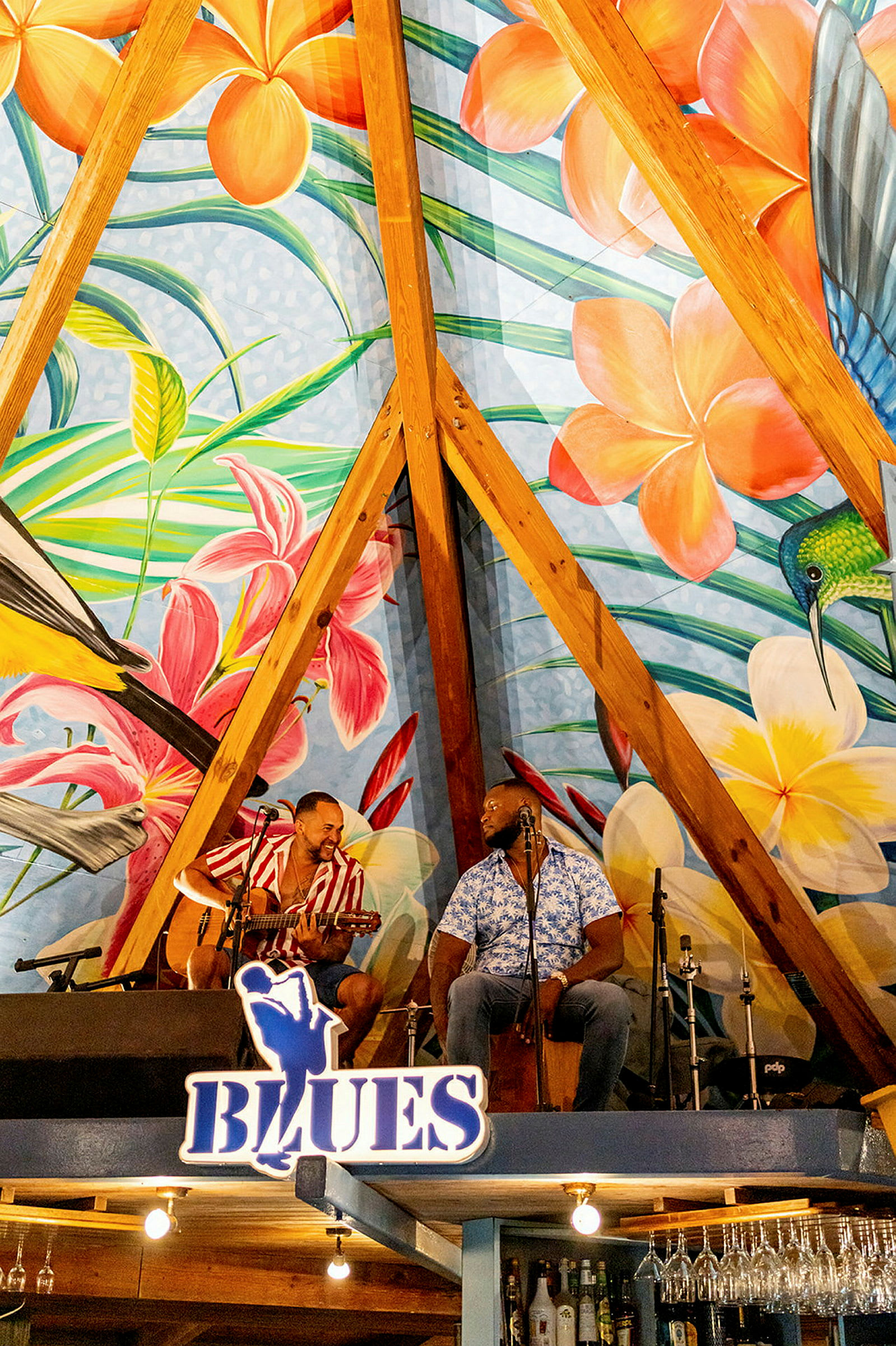 En färgglad mural med blommor och fåglar pryder väggen i en trästruktur ovanför en scen med musikinstrument. (AI-genererad text)