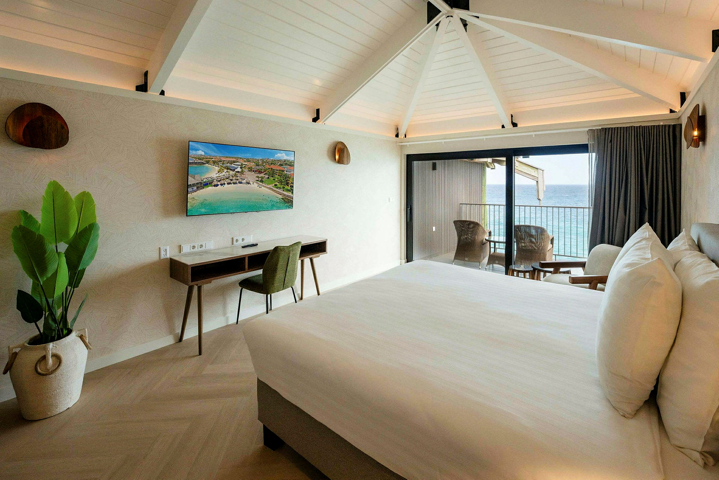 Ett hotellrum med en dubbelsäng, skrivbord, TV och balkong med utsikt över havet. (AI-genererad text)