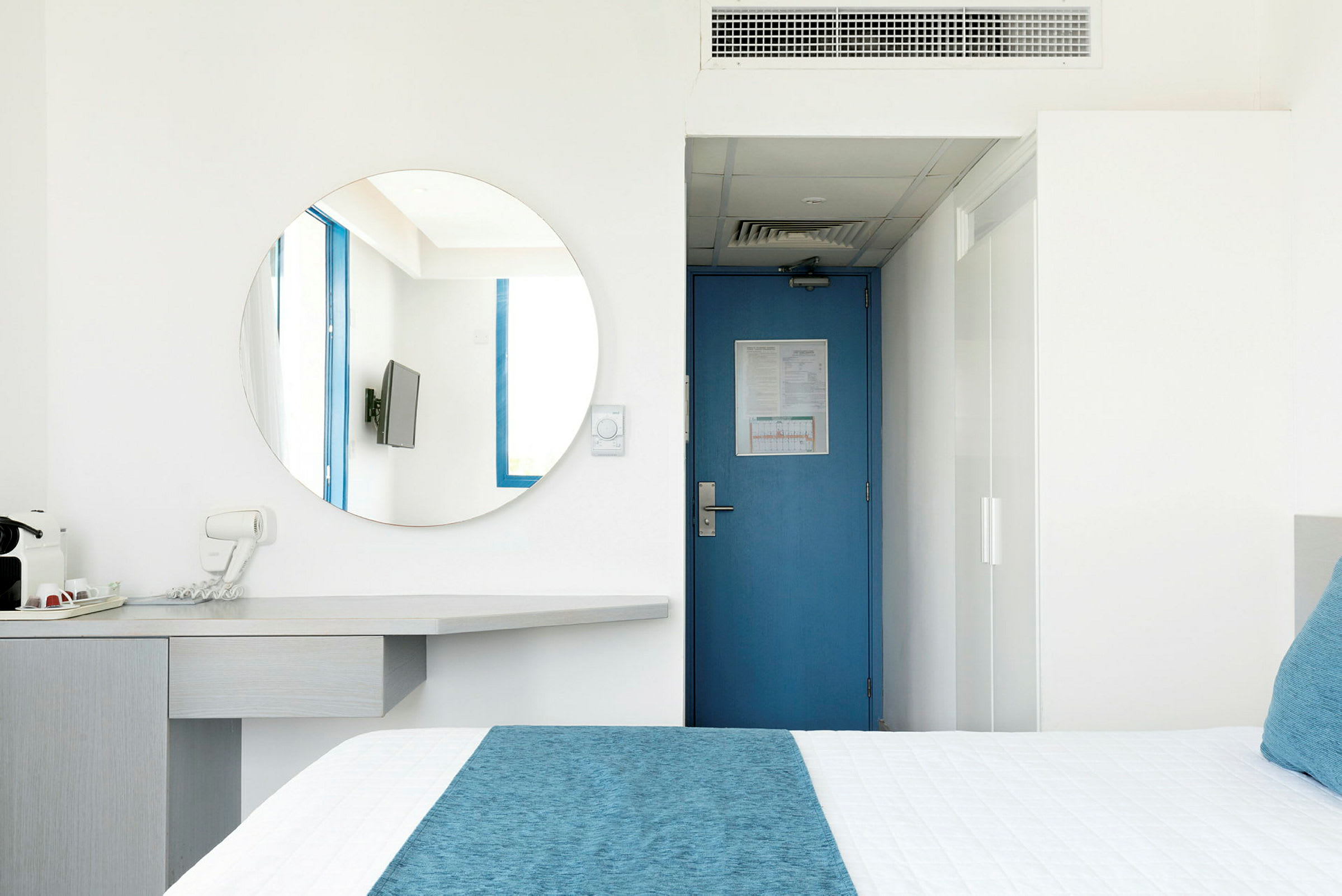 Ett hotellrum med en blå dörr, en rund spegel, en TV och en säng med blå detaljer. (AI-genererad text)