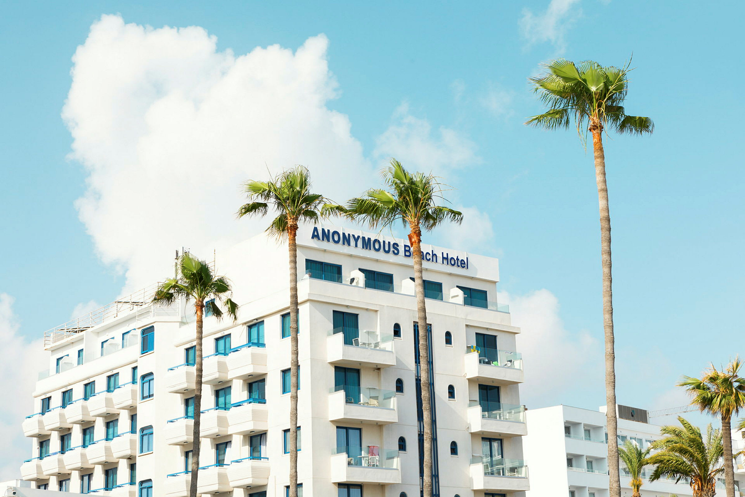 Ett hotell med namnet Anonymous Beach Hotel, vit fasad och blå fönster, omgivet av palmer. (AI-genererad text)