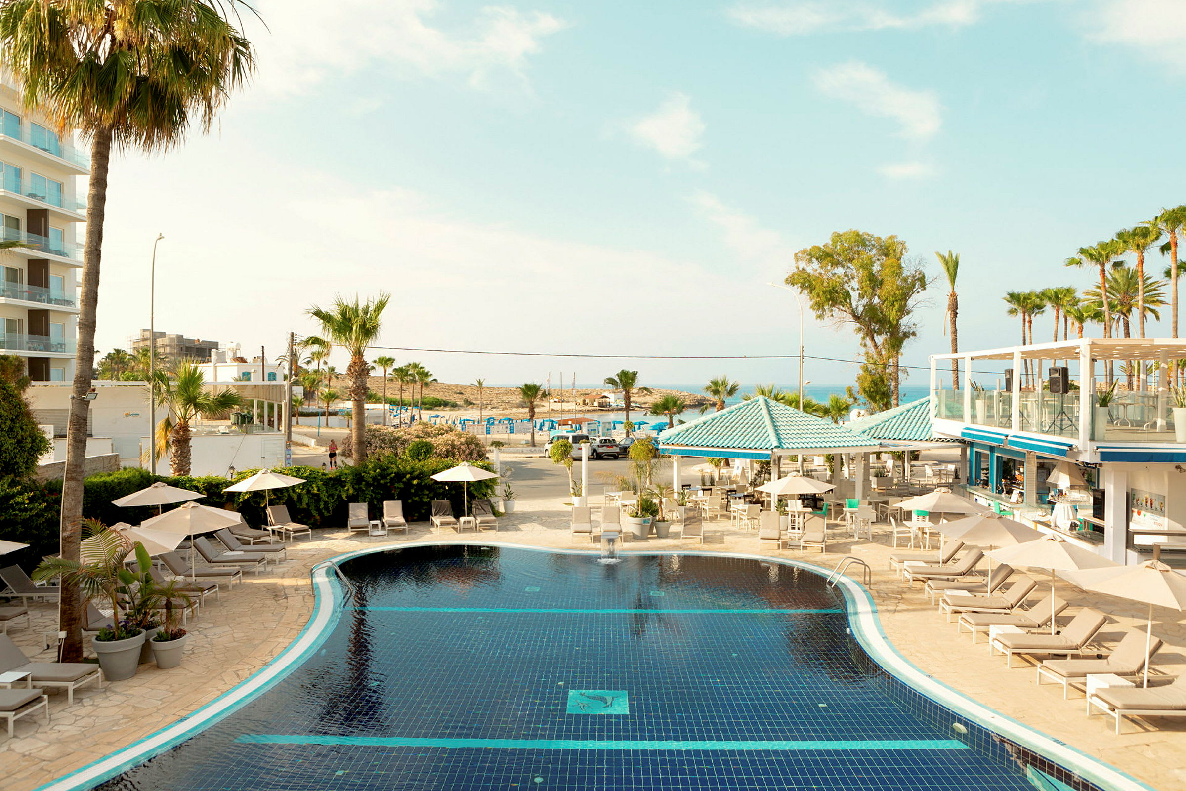 Hotellområde med pool, solstolar, parasoller och palmer, med strand och hav i bakgrunden. (AI-genererad text)