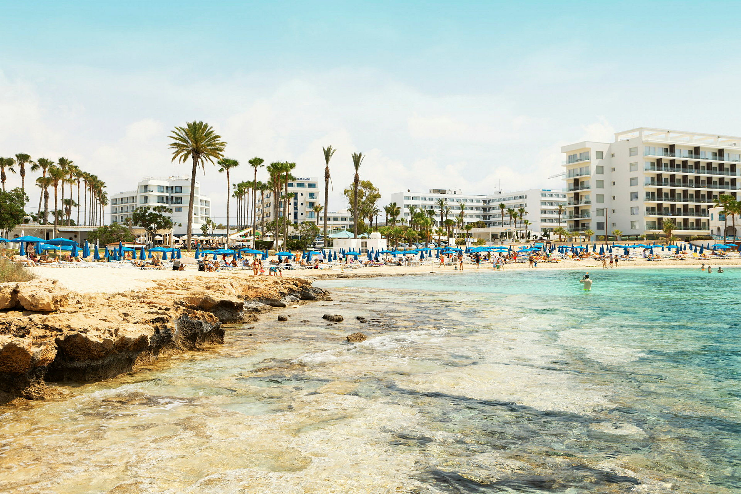 Strand med blå parasoller, palmer och hotellbyggnader i bakgrunden. (AI-genererad text)