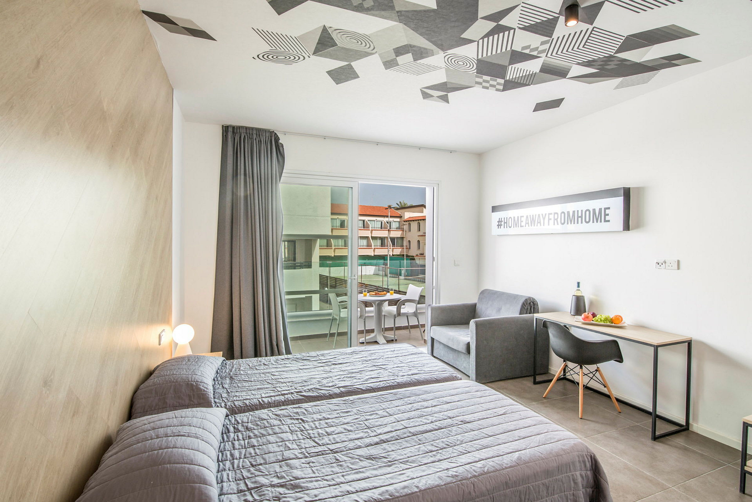 Ett hotellrum med två sängar, en soffa, ett skrivbord och en balkong med utsikt mot en byggnad. (AI-genererad text)