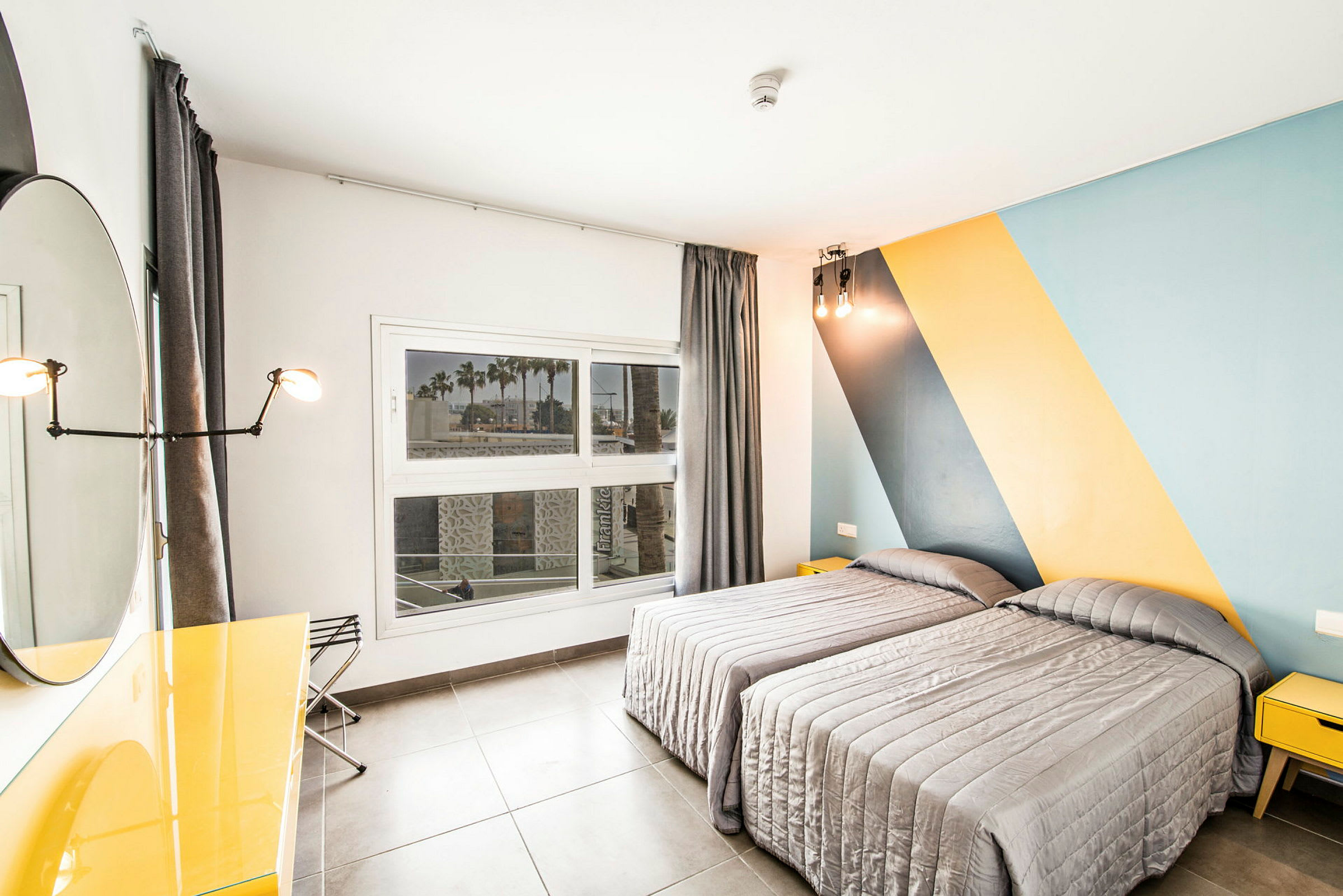 Ett sovrum med två enkelsängar med grå överkast. En stor fönsterruta med utsikt mot palmer och byggnader syns på väggen. Rummet har en modern, färgglad design med blå och gula diagonalpaneler. (AI-genererad text)